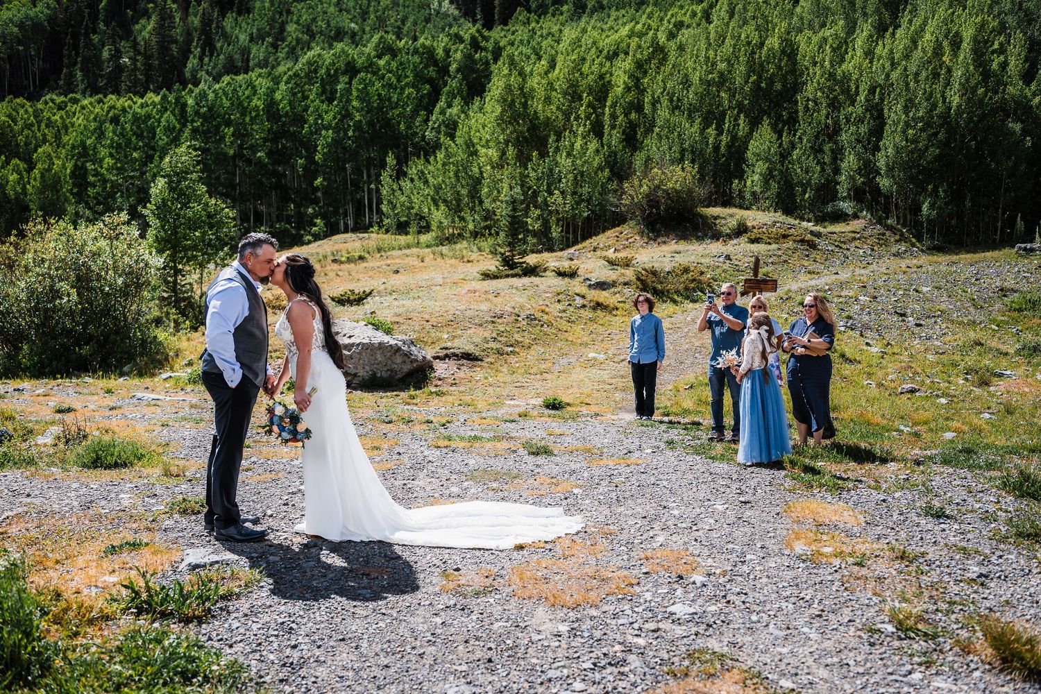 Bride and groom share a first kiss during an outdoor mountain elopement ceremony with guests on a sunny day.