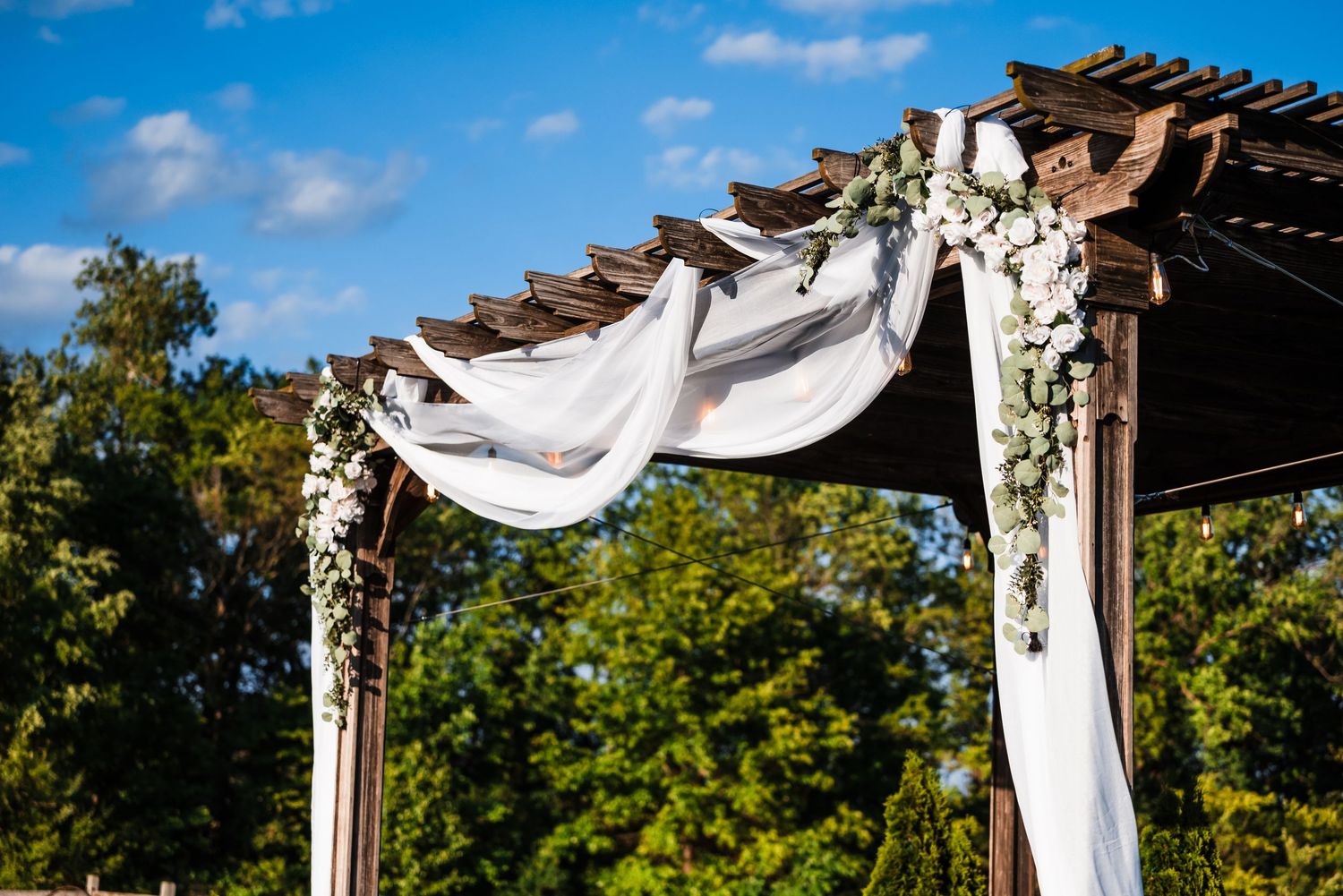 Elegant wooden wedding arch adorned with white draping fabric and fresh floral greenery under a bright blue sky.