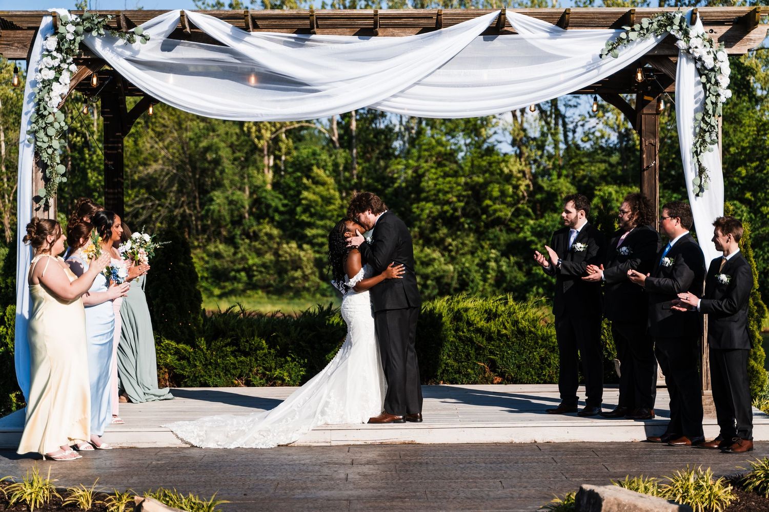 Bride and groom sharing first kiss under draped wedding arch with wedding party applauding.