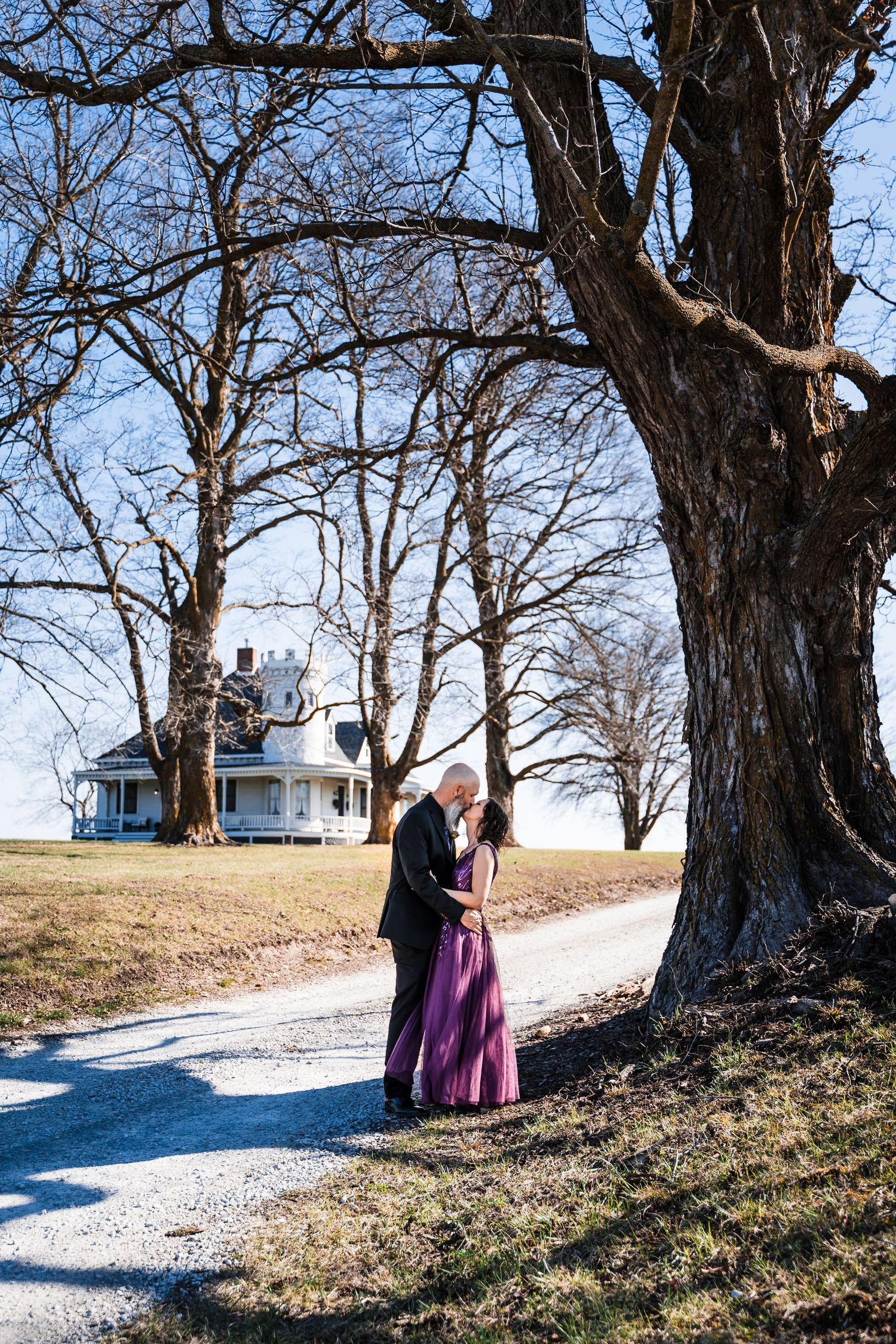 Wedding couple embracing under bare winter trees on gravel path with historic house in background.