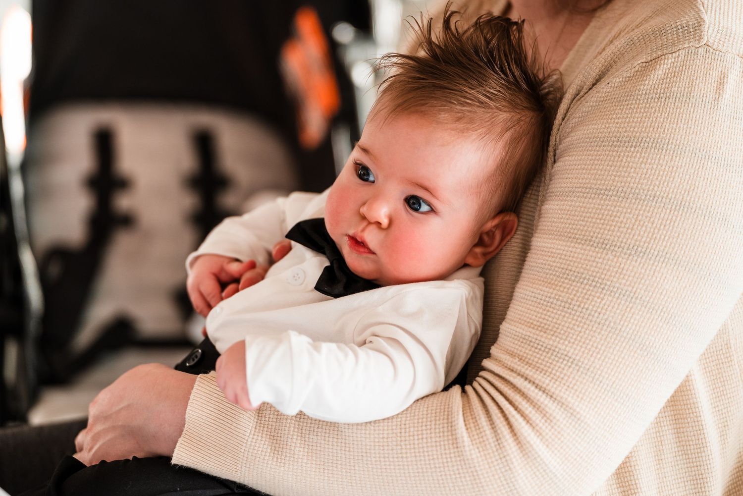 A cute baby with blue eyes being held by an adult in a cream sweater near a black stroller.