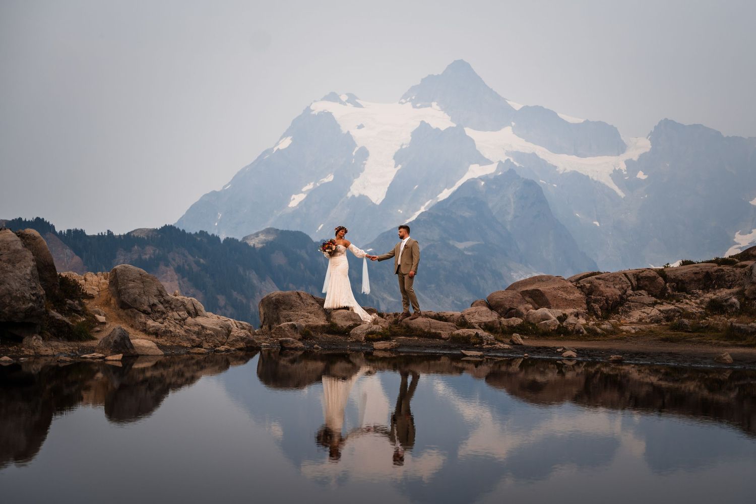 Two people in formal attire stand on rocky terrain near a mountain lake reflecting snow-capped peaks at sunset.
