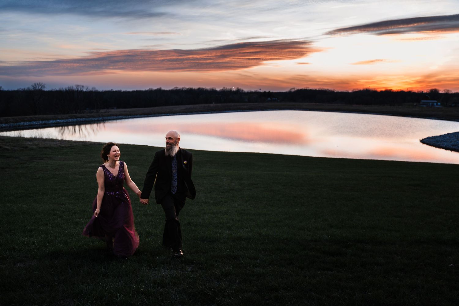 Bride and groom walk hand in hand beside a reflective pond at dramatic sunset.