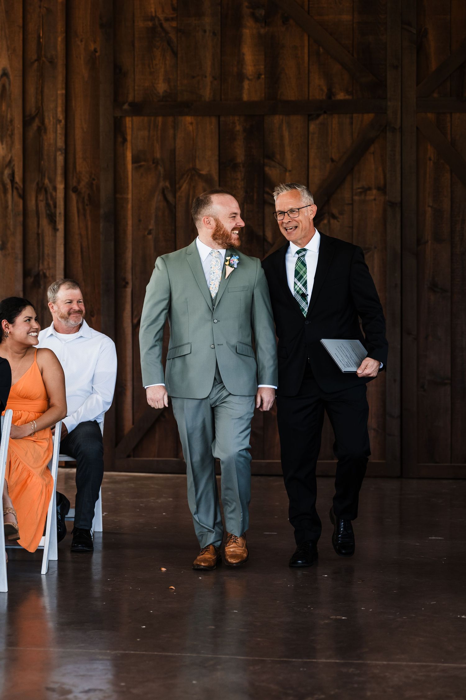 Groom in sage green suit walks down barn aisle with older gentleman in black suit at rustic wedding ceremony.