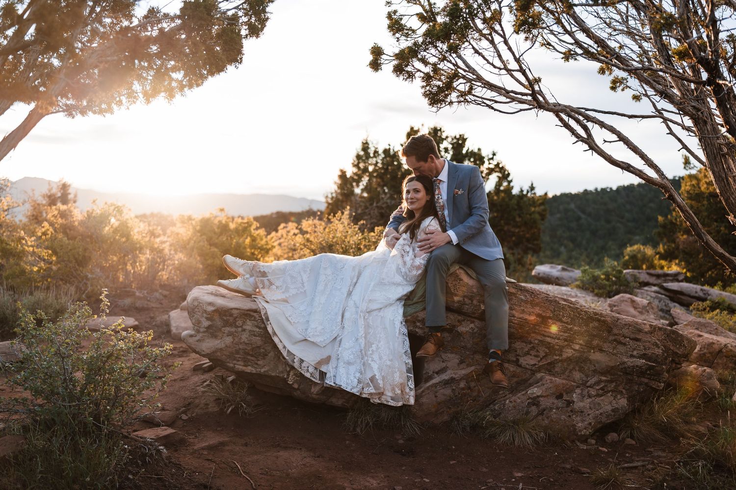 Groom kissing bride seated on a rock ledge surrounded by golden sunset light and desert landscape.
