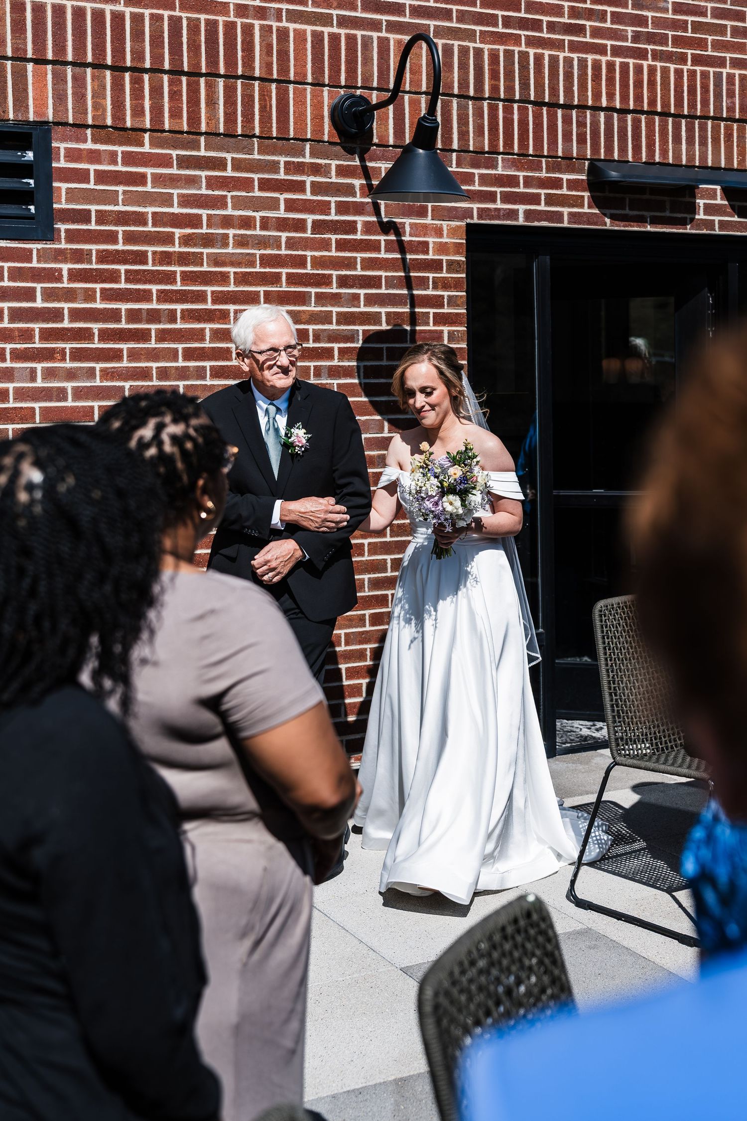 Bride walks down outdoor aisle with elderly father escort at sunny brick building wedding.
