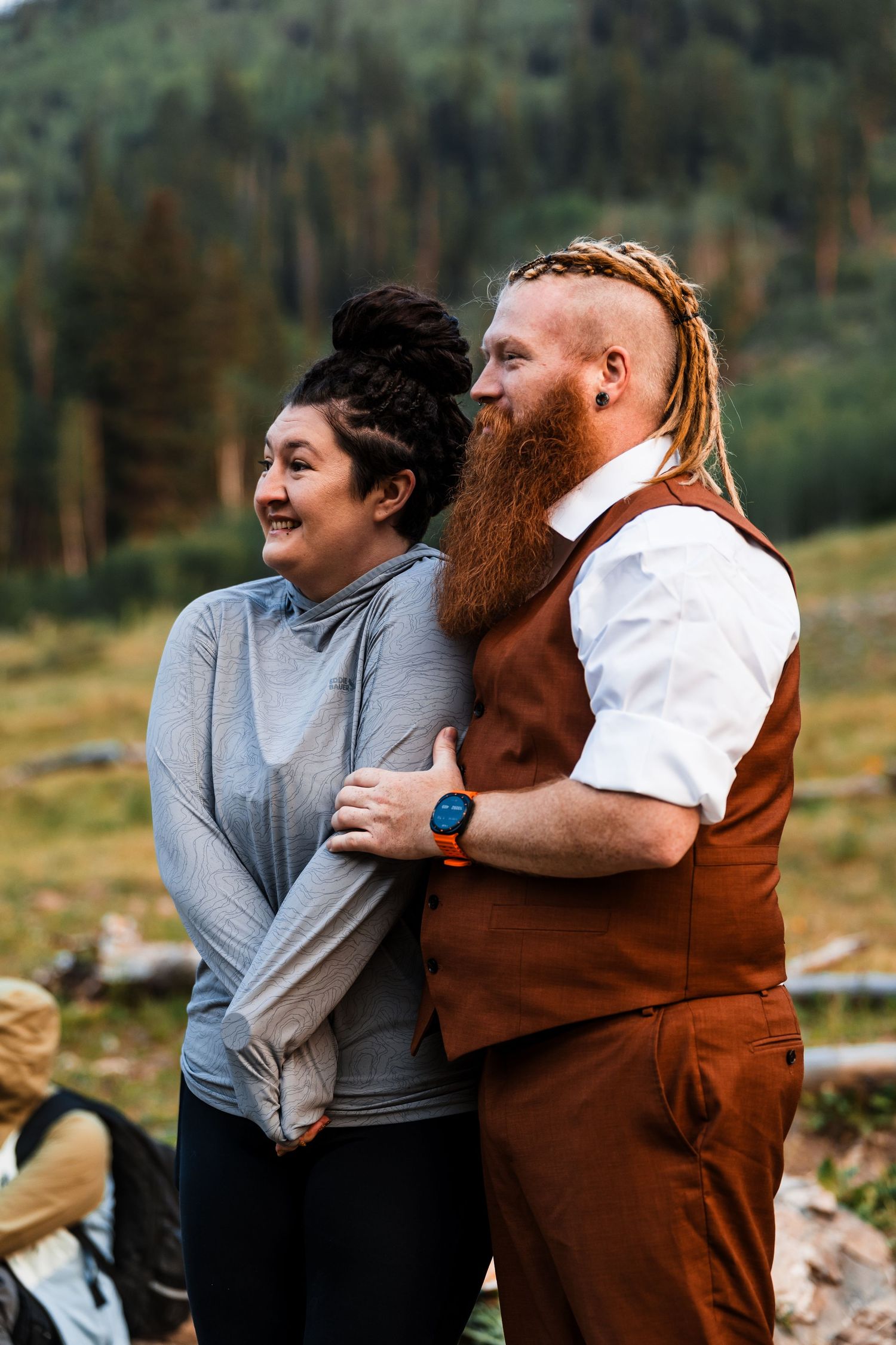 Couple embracing outdoors in mountain forest setting, man with red dreadlocks and beard wearing brown vest hugs smiling woman.