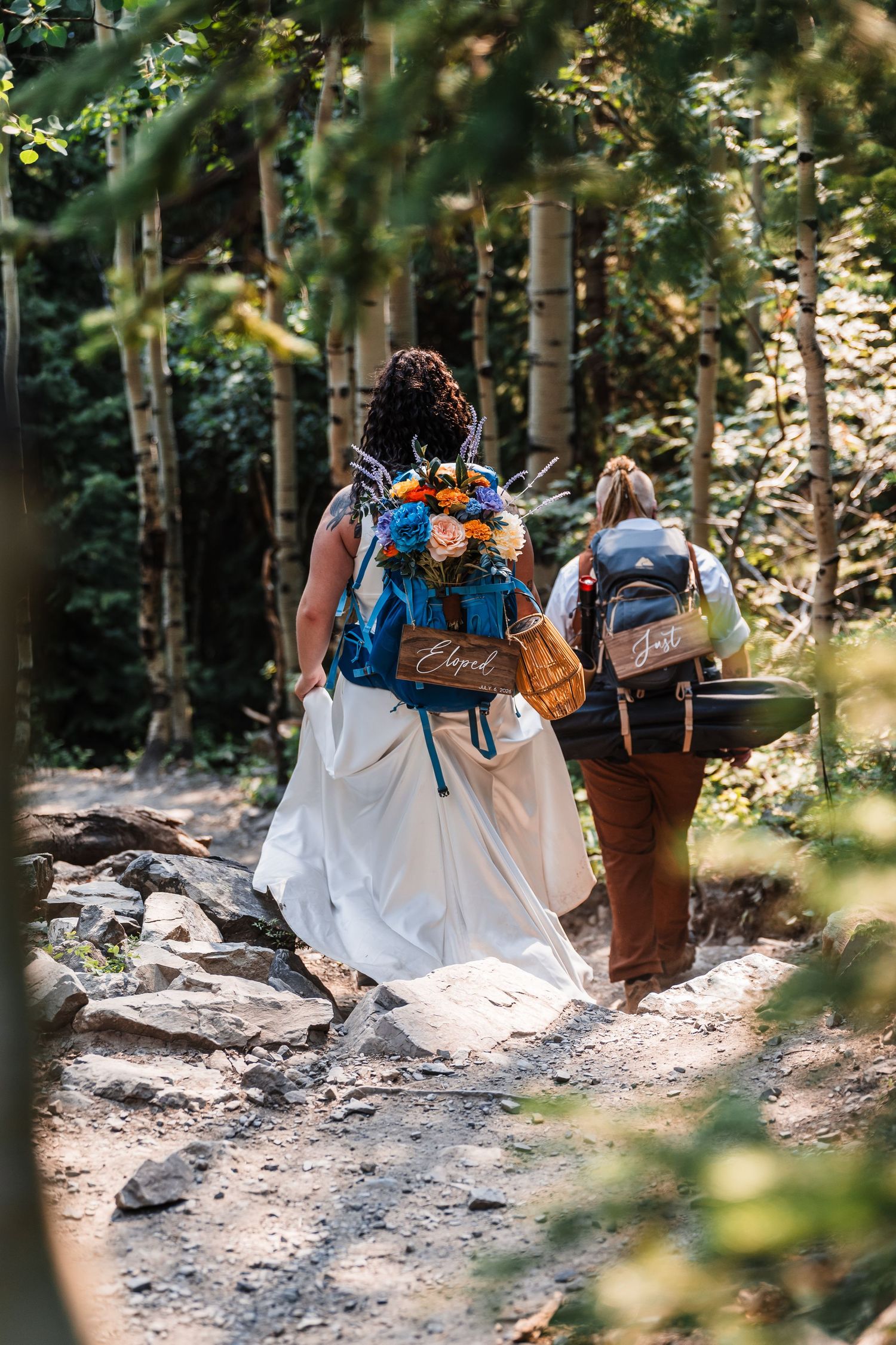 Bride in white dress and companion hike forest trail with backpacks adorned with wooden name signs and flowers.