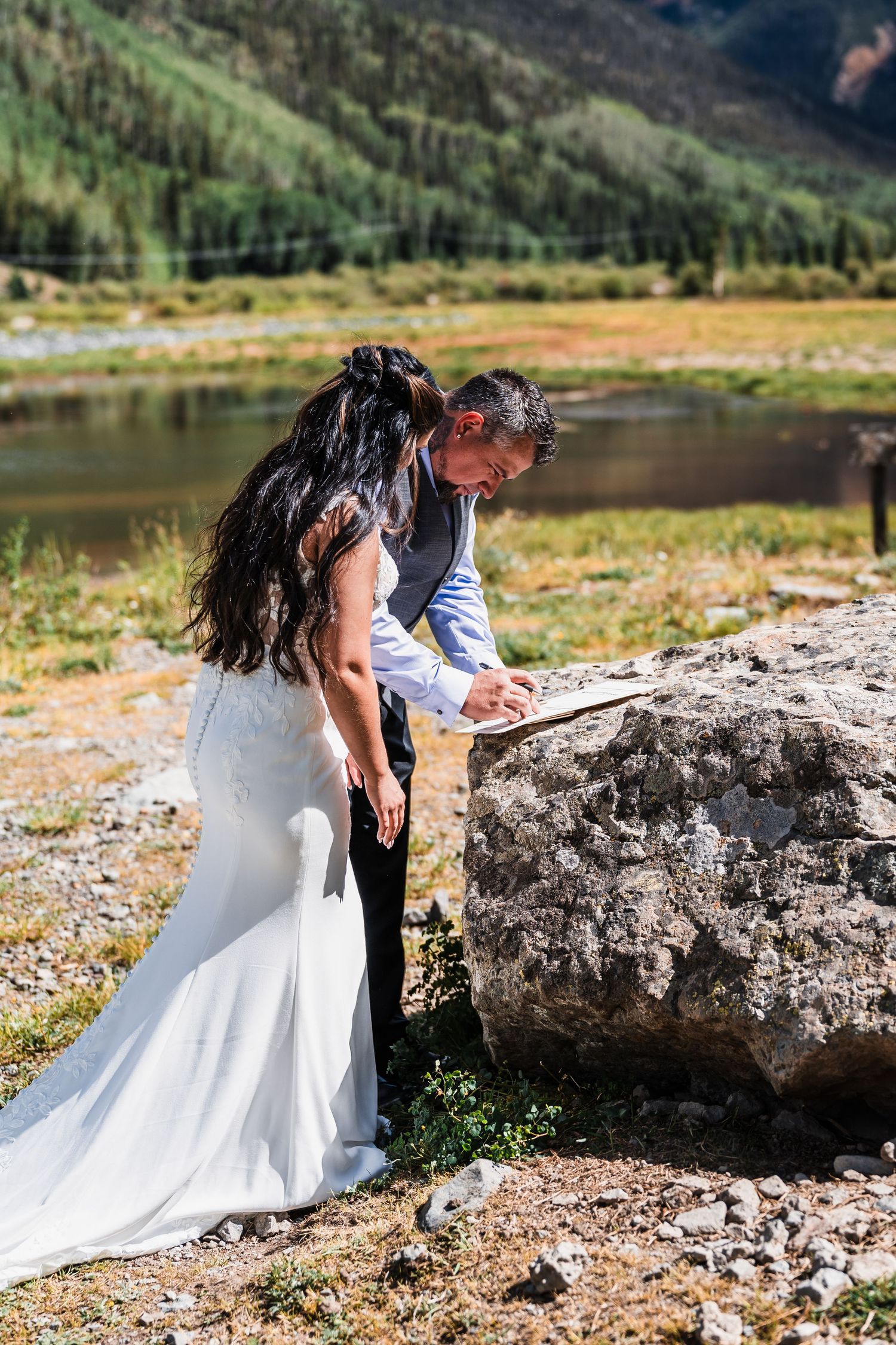 Bride and groom signing wedding documents on a rock by a mountain lake in Colorado.