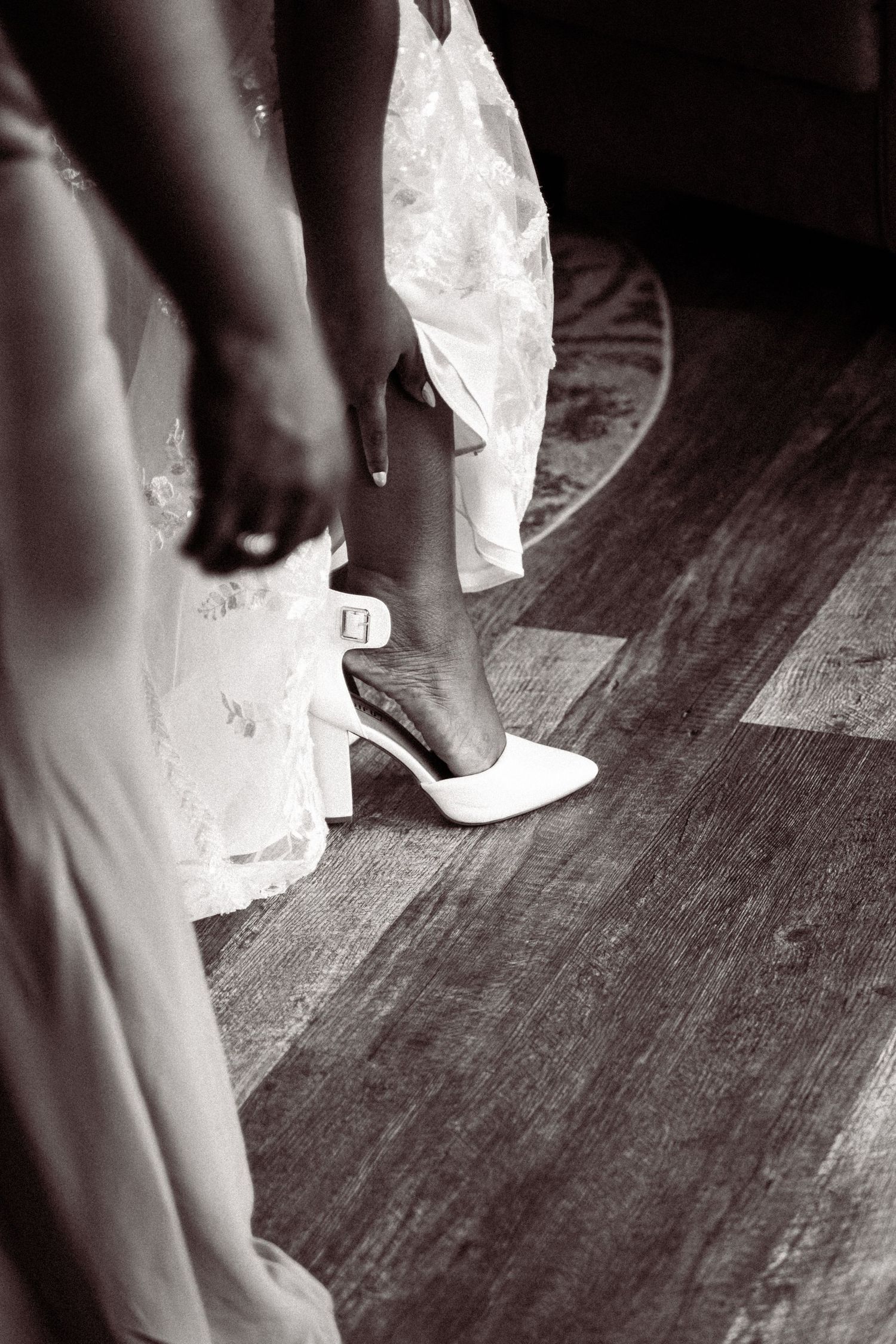 Black and white photo of a bride putting on white heeled wedding shoes with ankle strap on wooden floor.