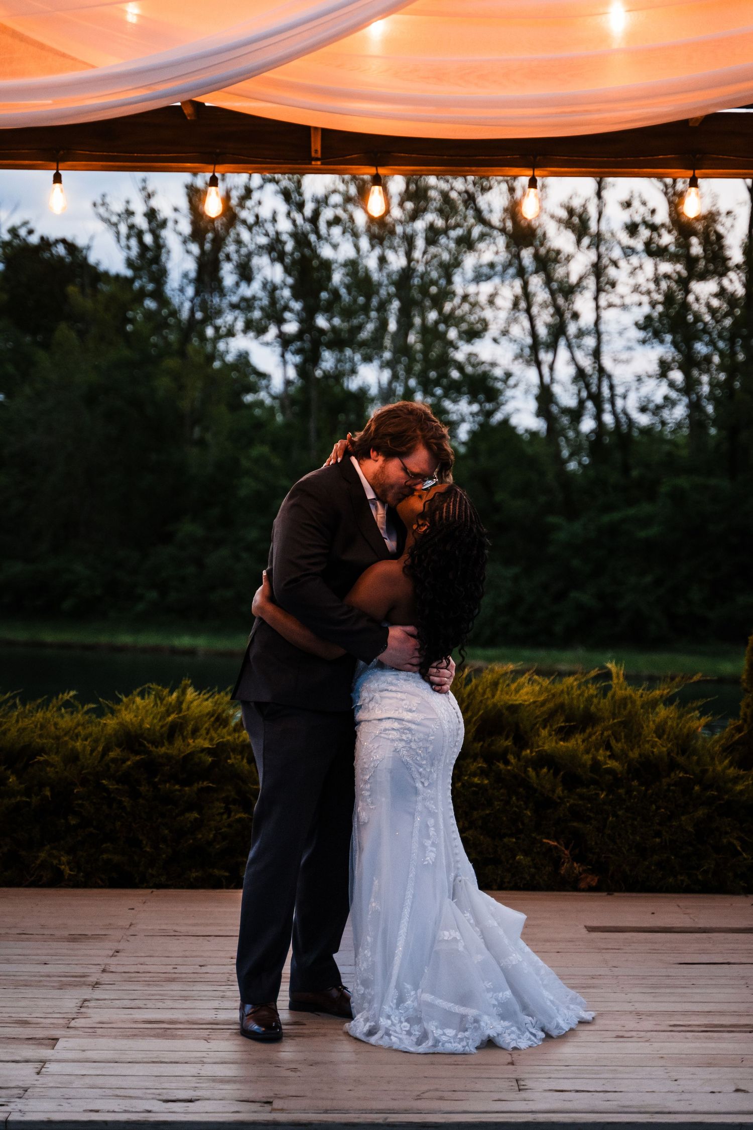 Newlyweds share romantic kiss on outdoor dance floor under warm string lights at twilight reception.