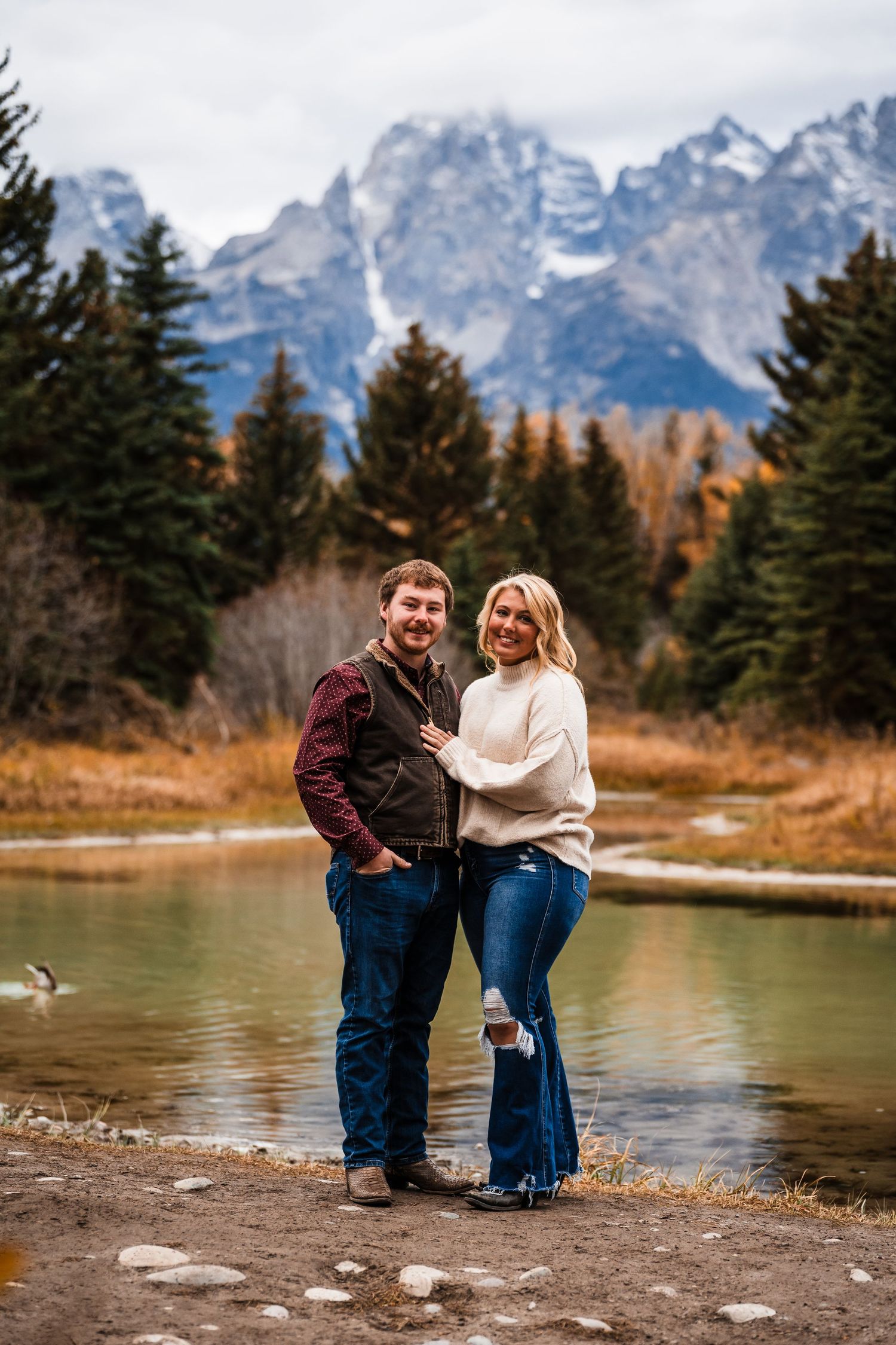 A couple stands together by a lake with the majestic Grand Teton mountains rising in the background on an autumn day.