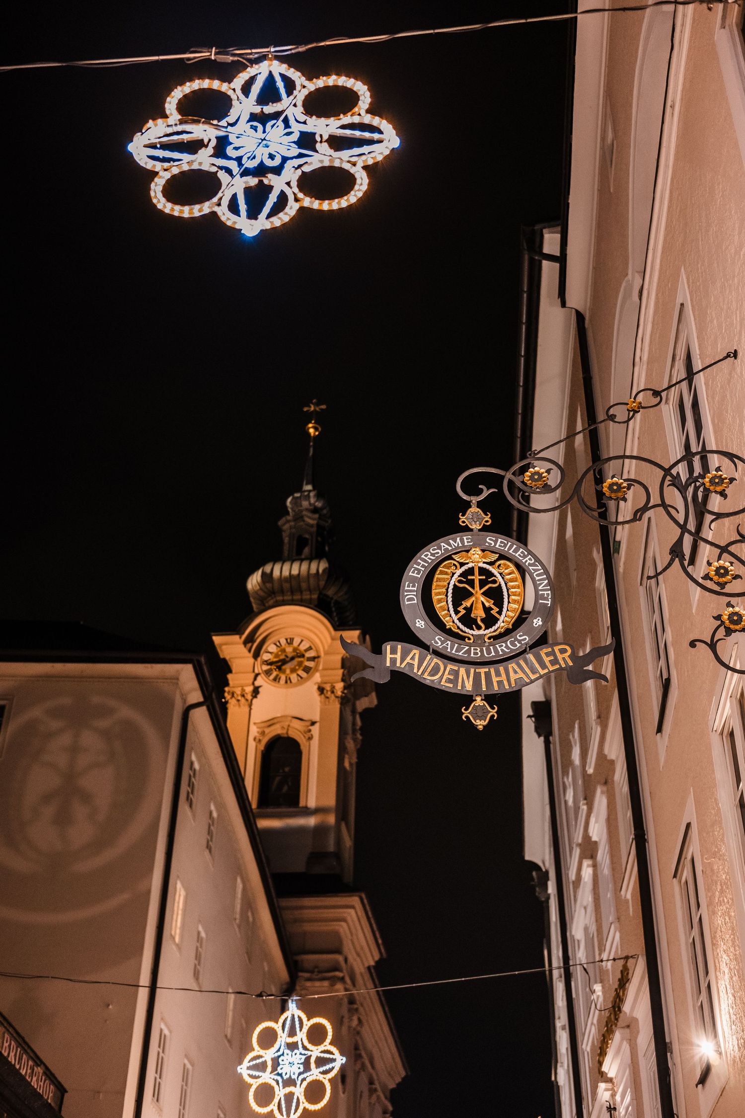 Decorative illuminated signs and ornate light fixtures hanging between historic buildings at night in a European city.