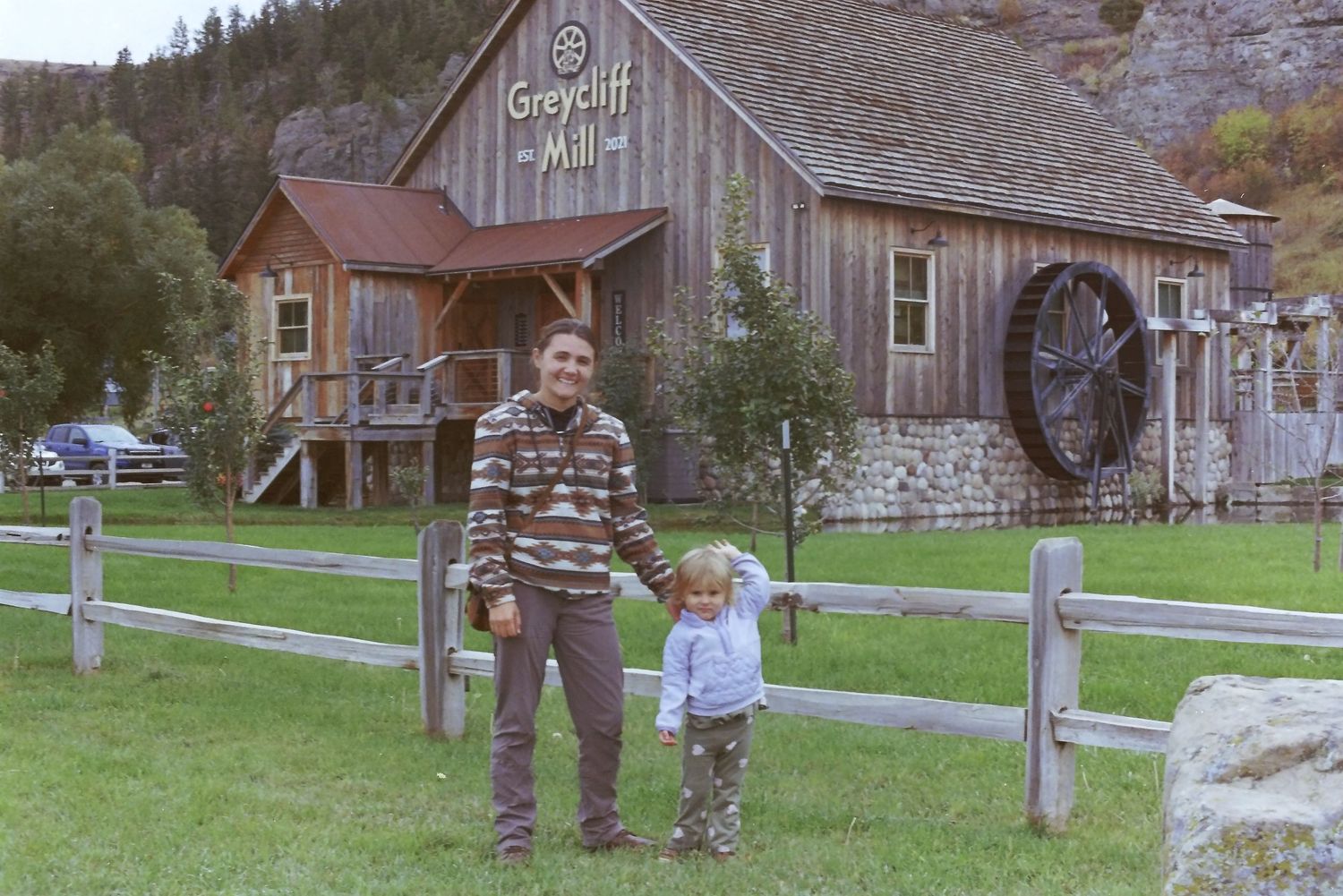 A rustic wooden barn with a water wheel serves as backdrop to people standing by a wooden fence on grassy grounds.