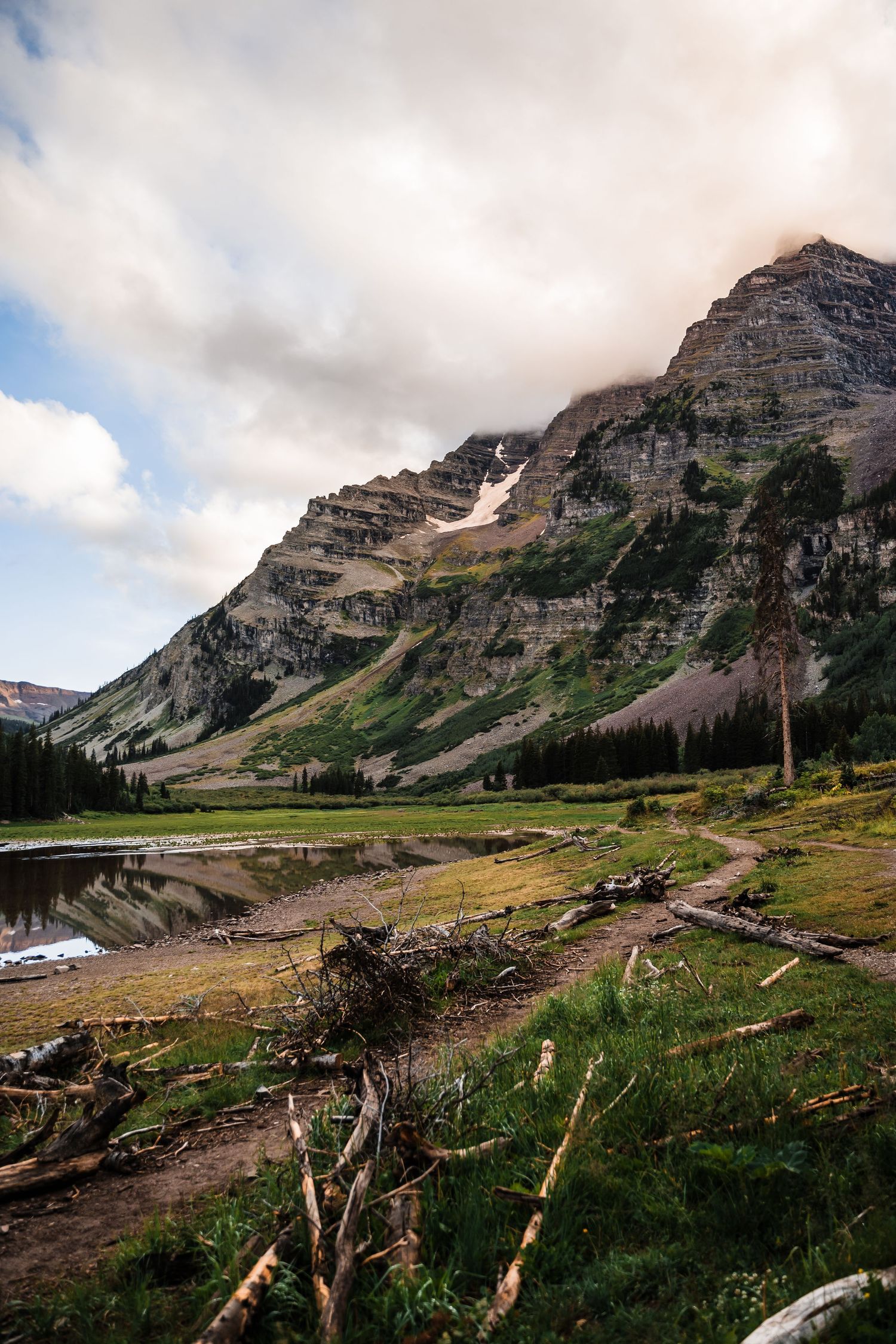 Dramatic mountain landscape with layered rock formations and alpine meadow reflected in a small lake under cloudy skies.