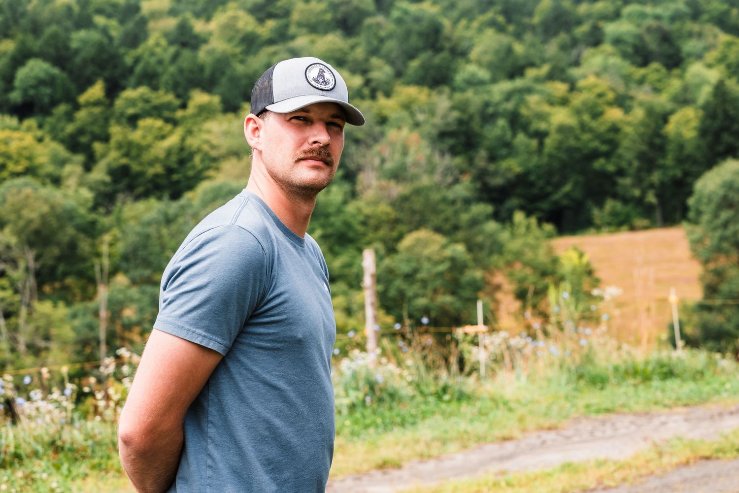 A person in a gray t-shirt and trucker hat stands outdoors against a backdrop of lush green hills.