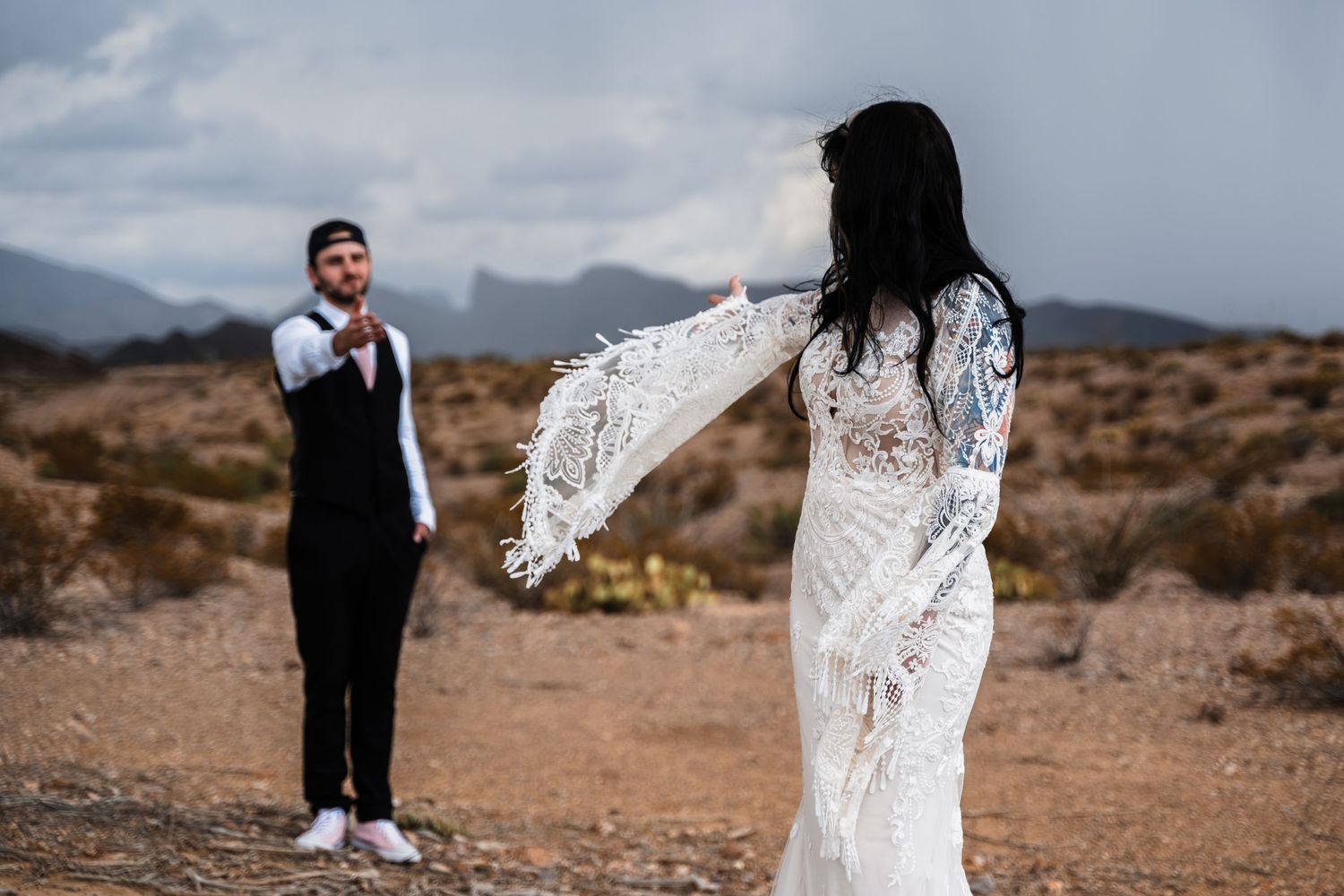 Bride in lace dress with flowing veil against desert mountain backdrop.