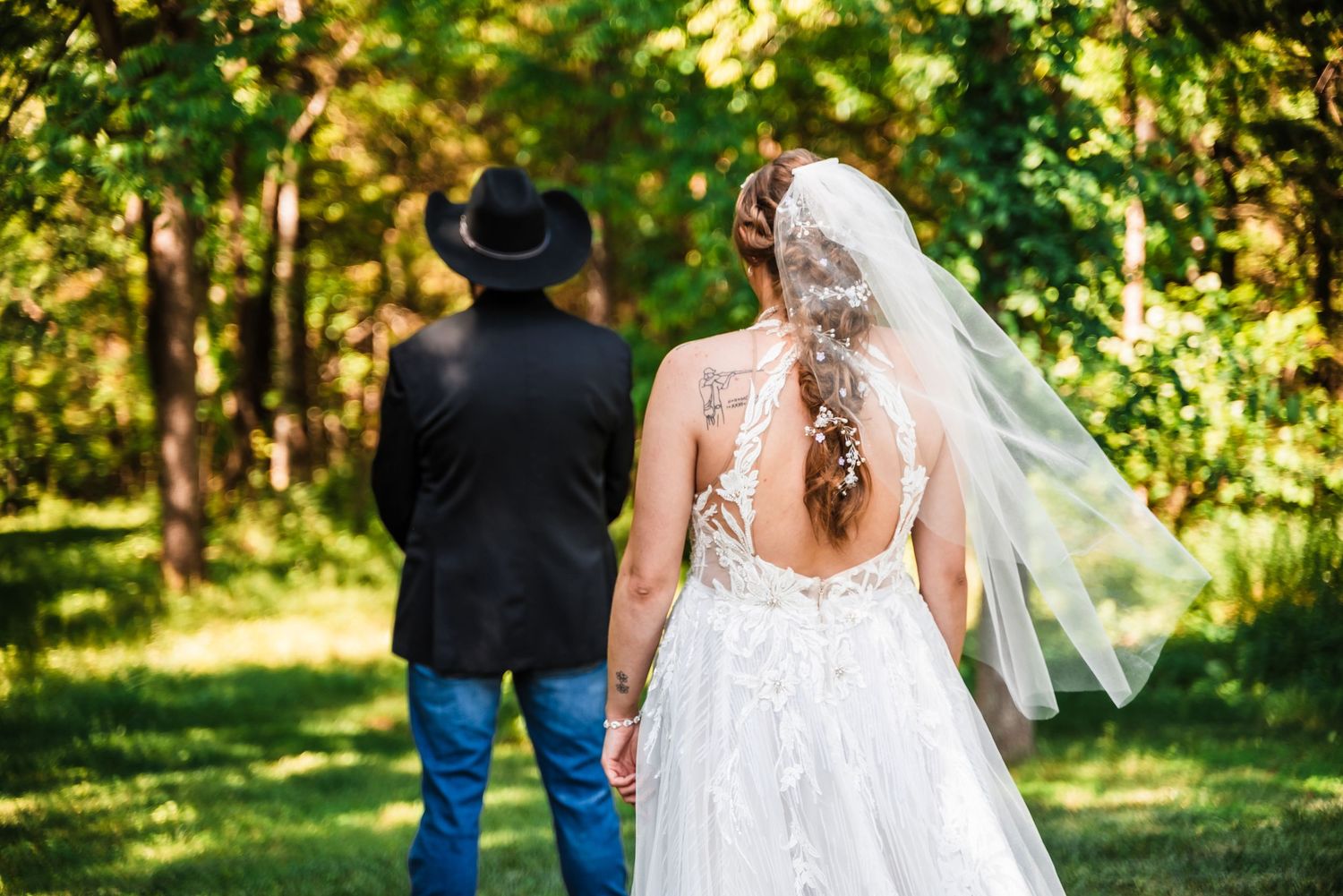 A rear view of a bride in a detailed lace gown with flowing veil standing in a sunlit garden setting.