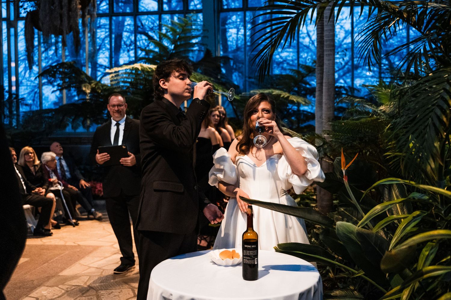 Newlyweds celebrate with a champagne toast during an indoor wedding ceremony surrounded by tropical plants.