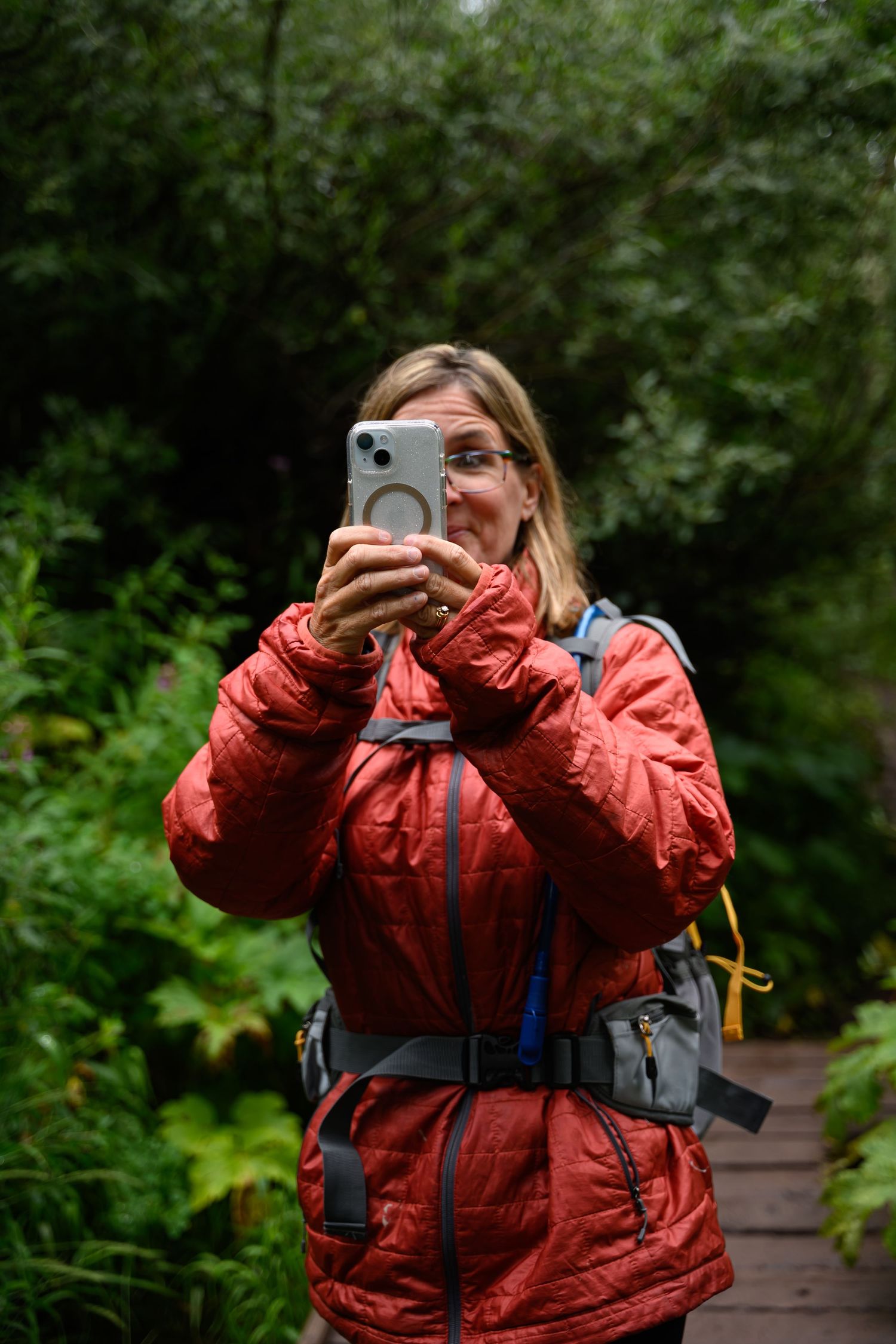 Hiker in red puffy jacket takes photo with phone on nature trail.