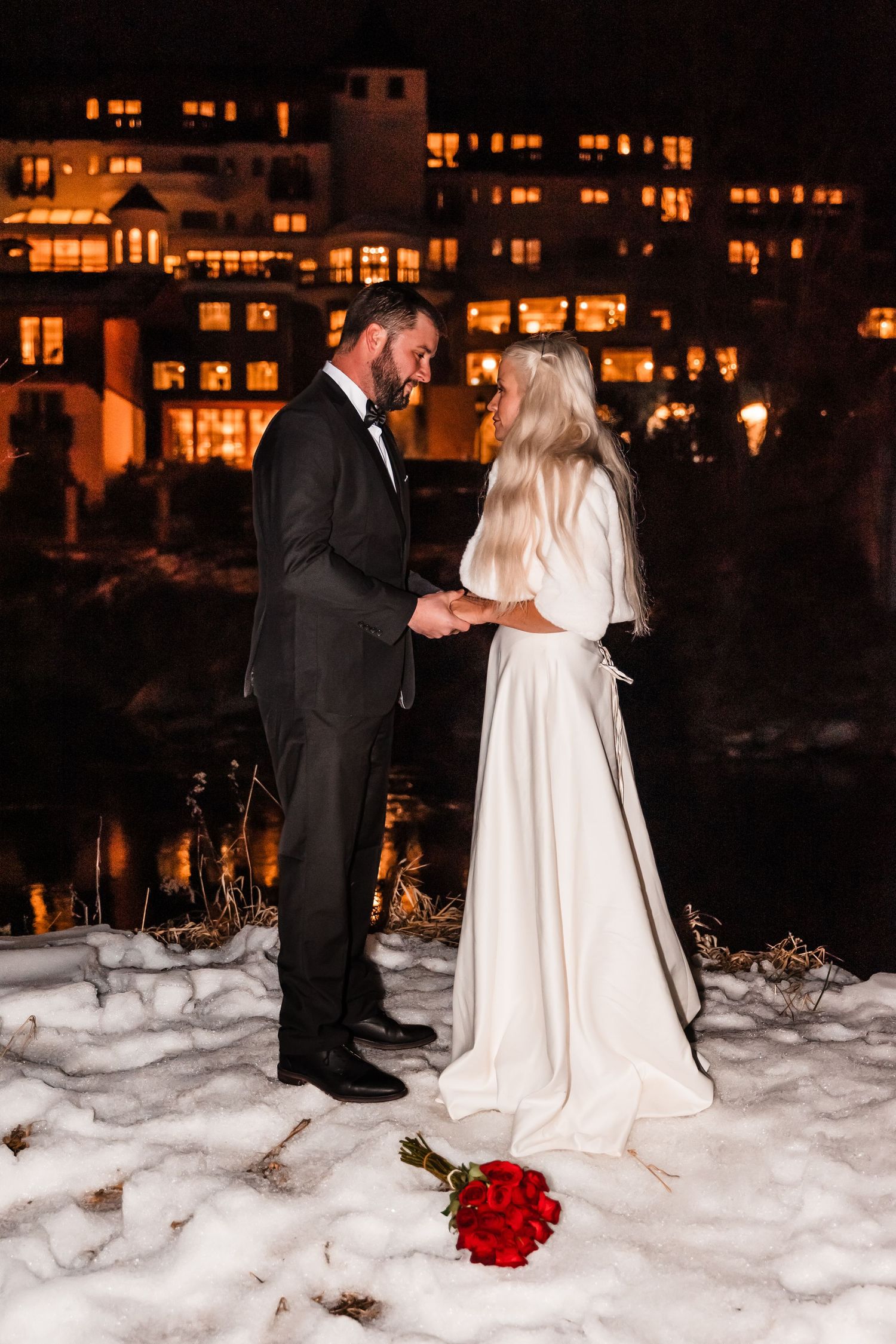 A nighttime winter wedding scene with glowing buildings in background and a red rose on snowy ground.