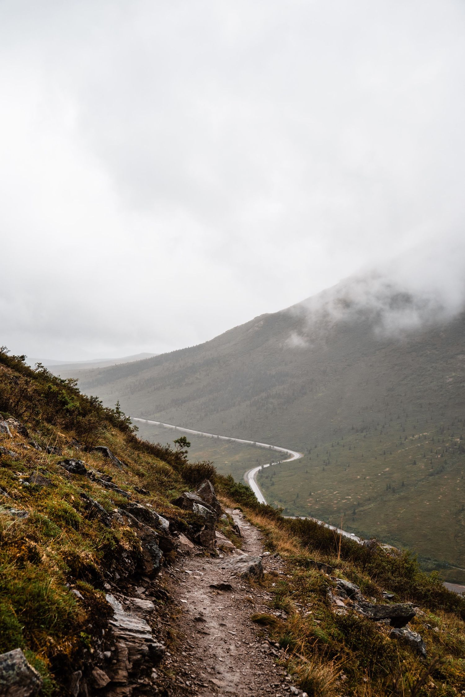 A misty mountain trail curves alongside a winding road below in a dramatic mountain landscape.