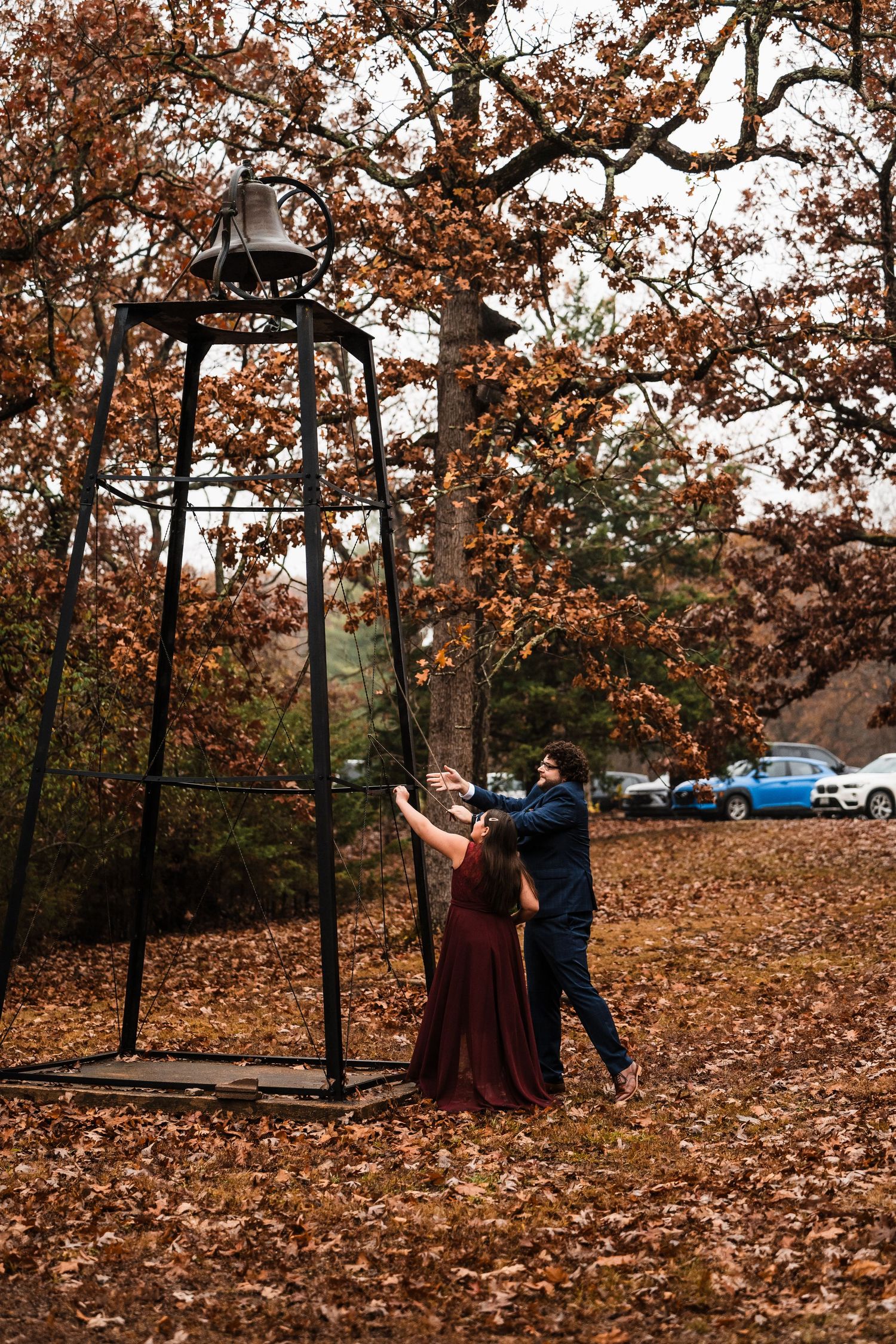 Autumn wedding photo with couple ringing large bell outdoors among fall foliage.