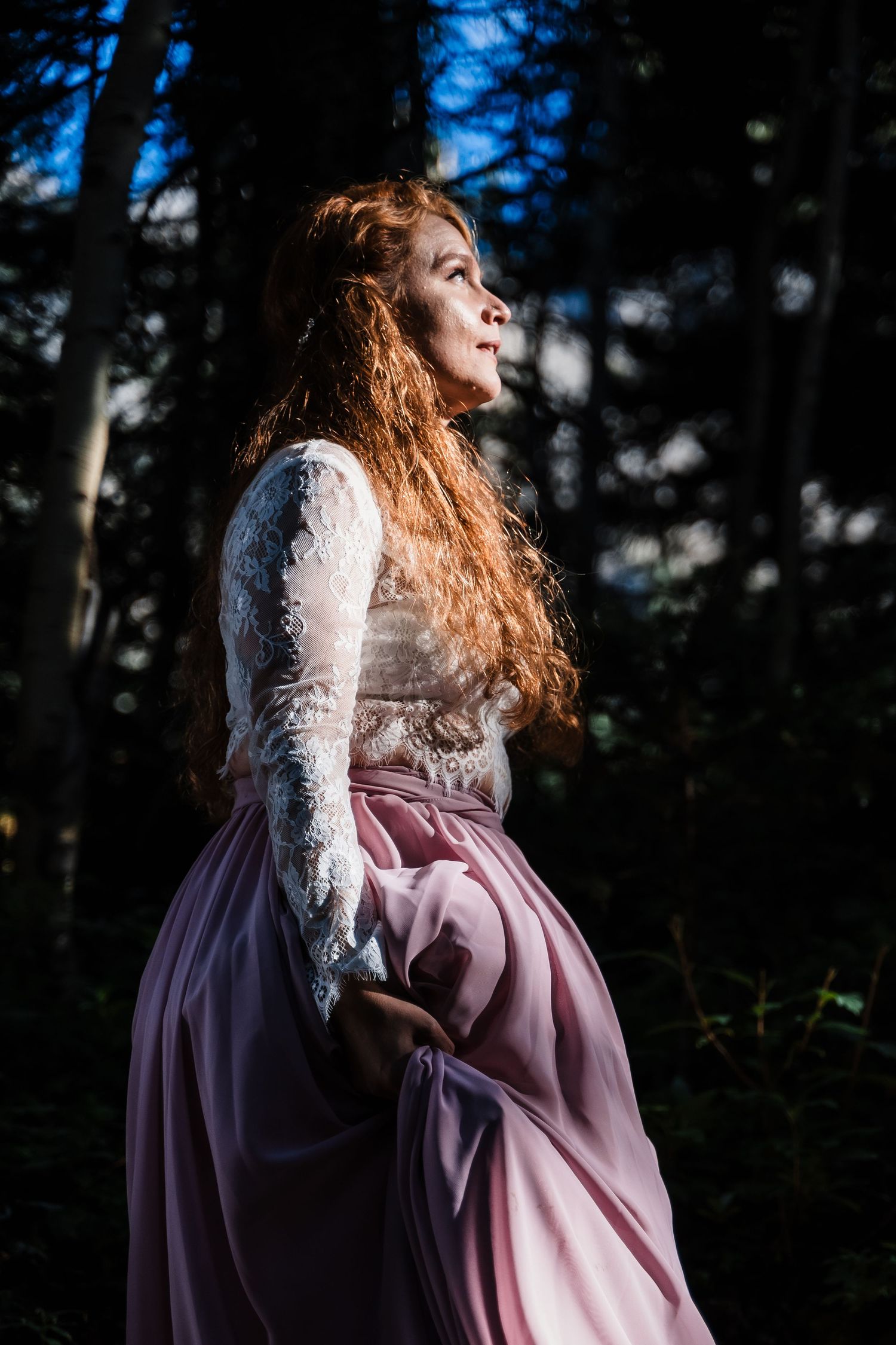 Figure in vintage-style dress poses dramatically in sunlit forest setting.