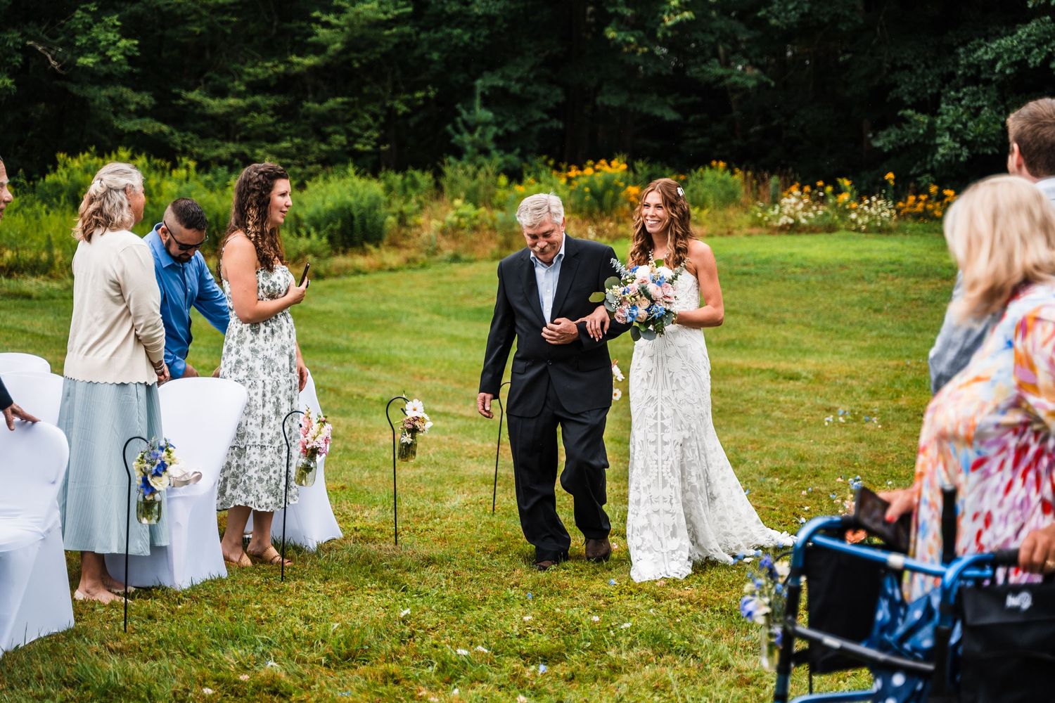 Wedding guests and family gather on a lawn with colorful flowers as a bride walks down the aisle.