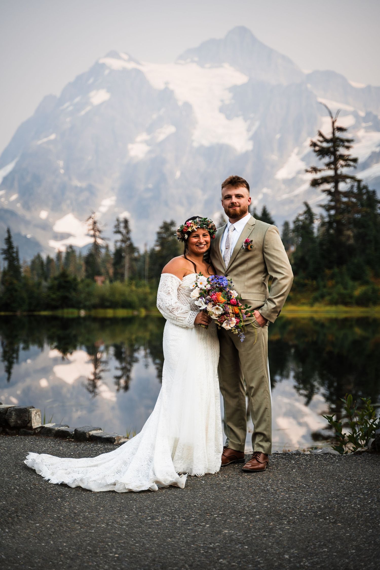 A couple poses for romantic wedding photos beside a reflective lake with snow-capped mountains behind them.