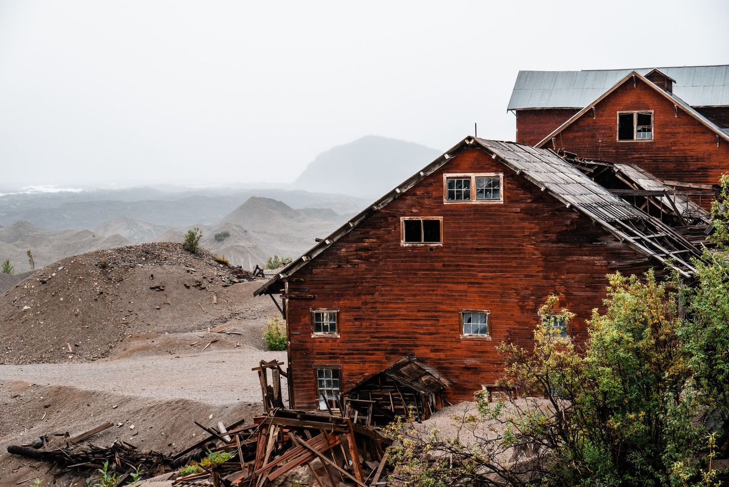 A weathered red wooden mining building perches on a rocky mountainside with misty peaks in the distance.