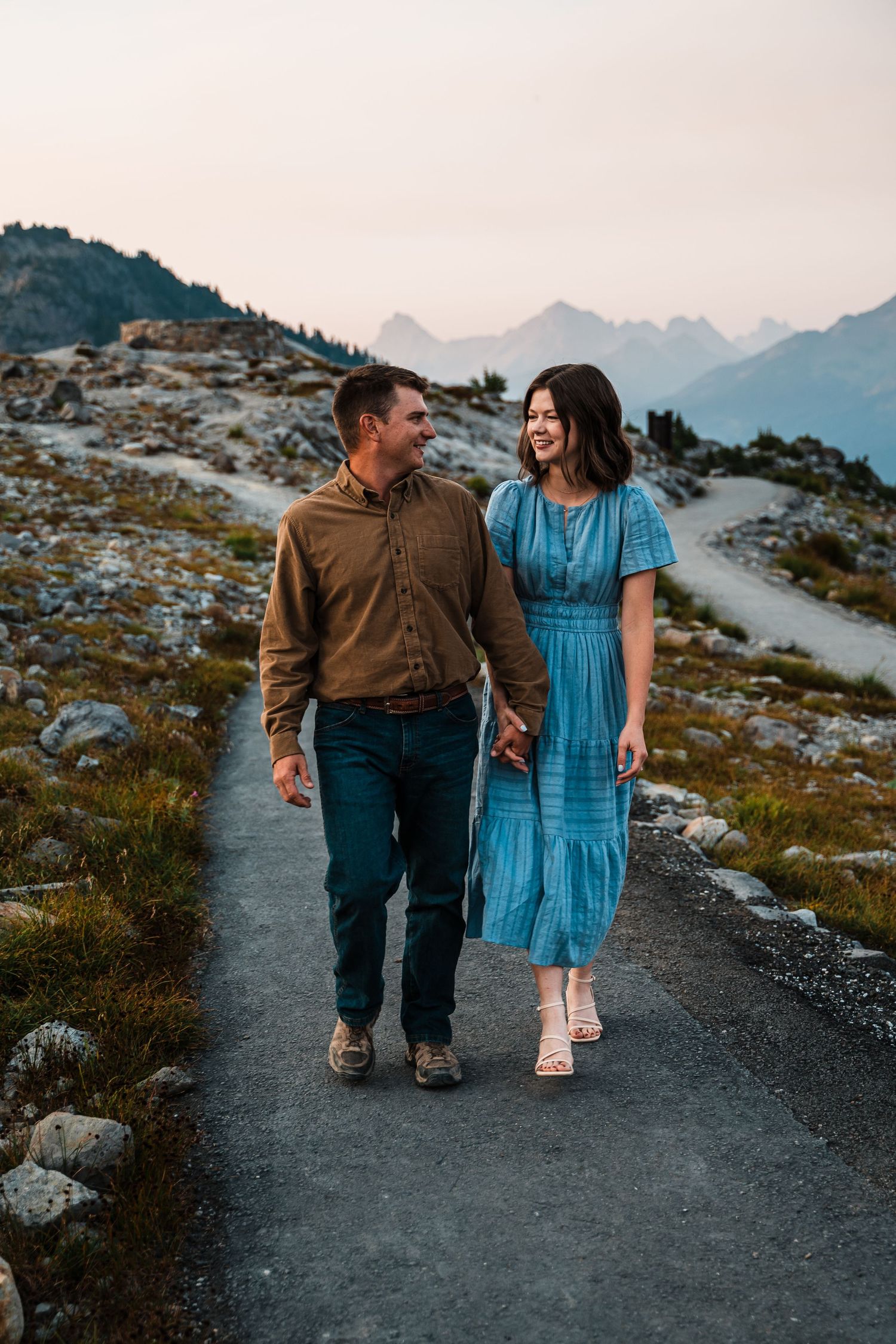 A couple walks hand in hand along a mountain trail at sunset with rugged peaks in the background.