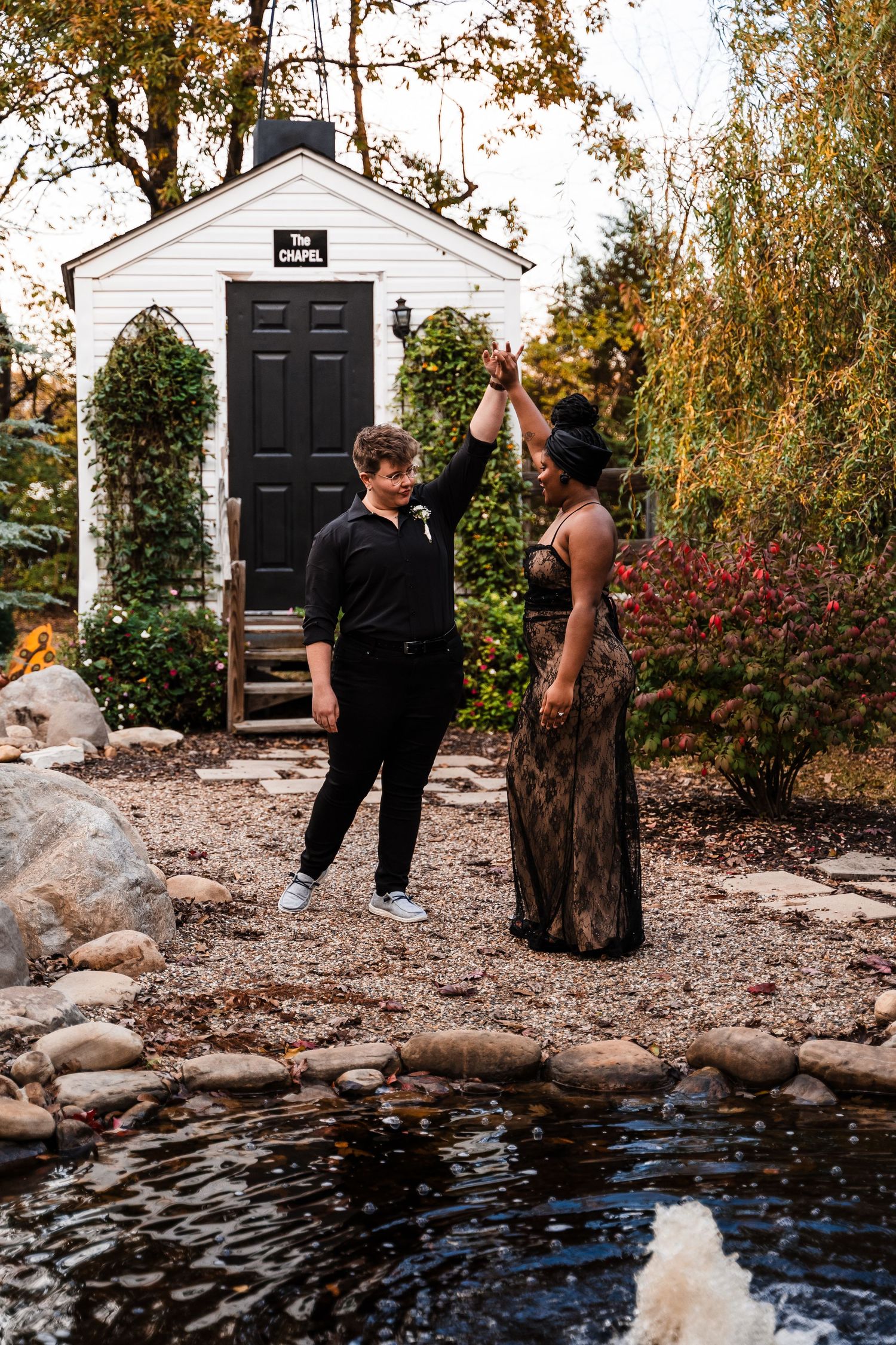 A couple celebrates together in front of a small white chapel with black door beside a decorative pond during autumn.