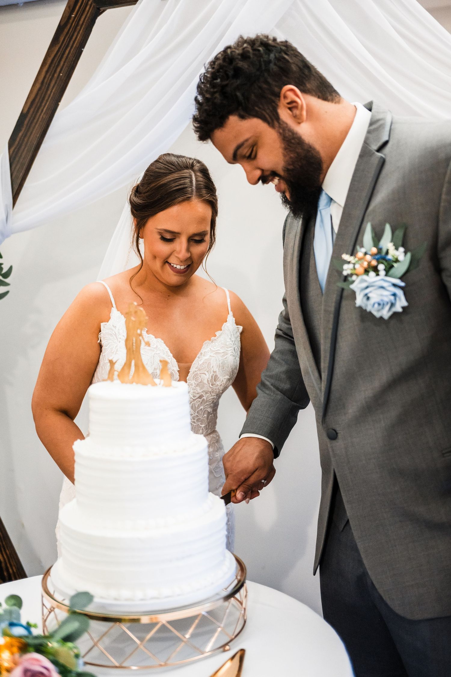 A newly married couple shares a sweet moment cutting their elegant white tiered wedding cake together beneath white draping.
