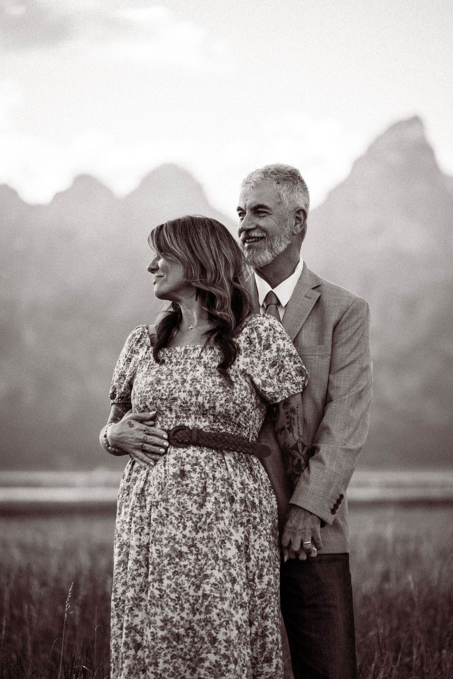 A couple embraces in a field at sunset, with the woman wearing a floral dress and the man in formal attire.