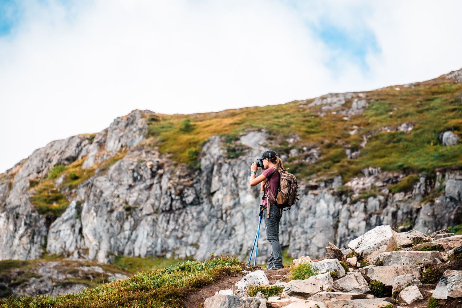 A hiker stands at the edge of a rocky cliff with mountain views and blue skies in the background.
