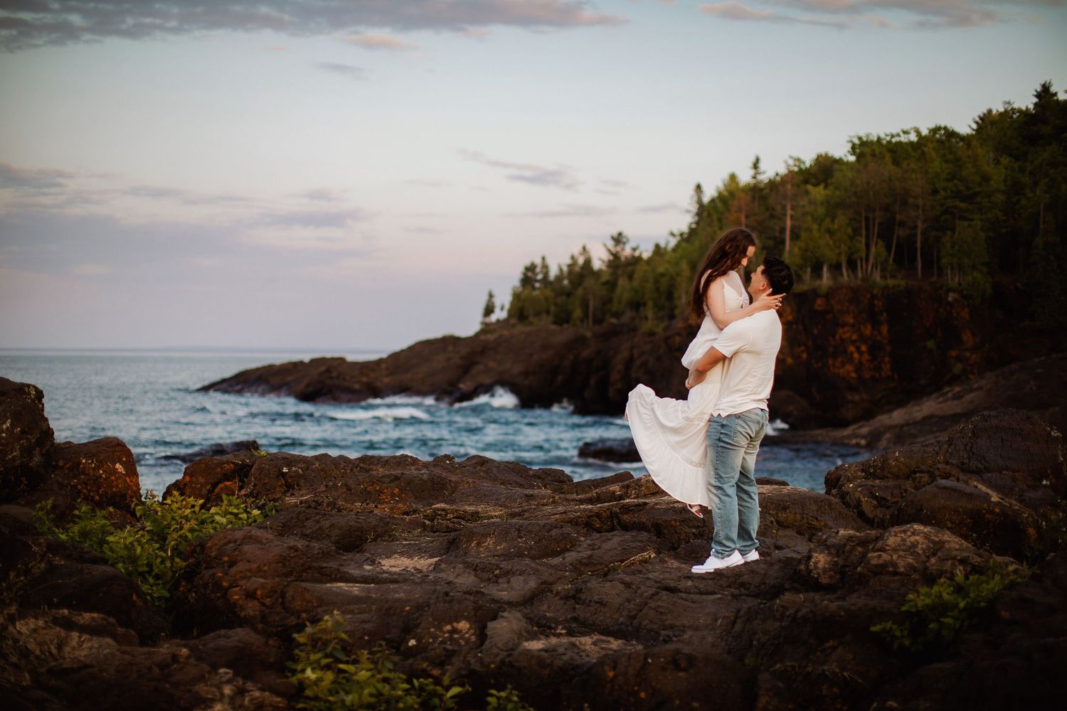 Engagement pictures on black rocks in Marquette Michigan during golden hour. Guy pick up girl with a leg pop on the Lake Superior shore
