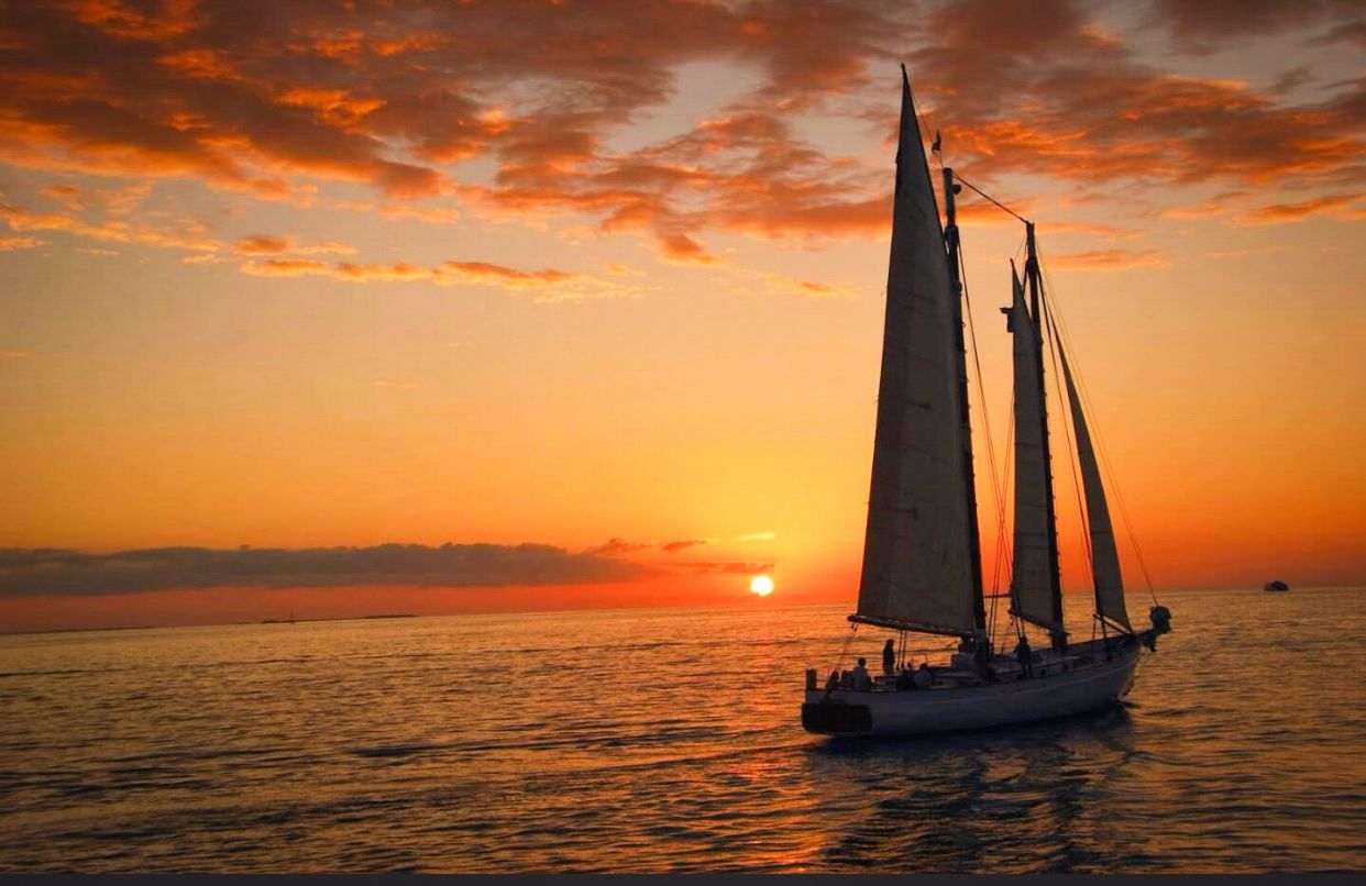 White sailboat sailing into breathtaking orange and red sunset over calm ocean waters.