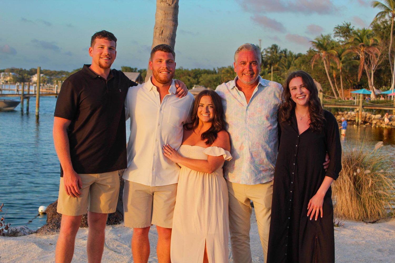 Family of five posing together on a tropical beach at sunset with palm trees and marina in background.