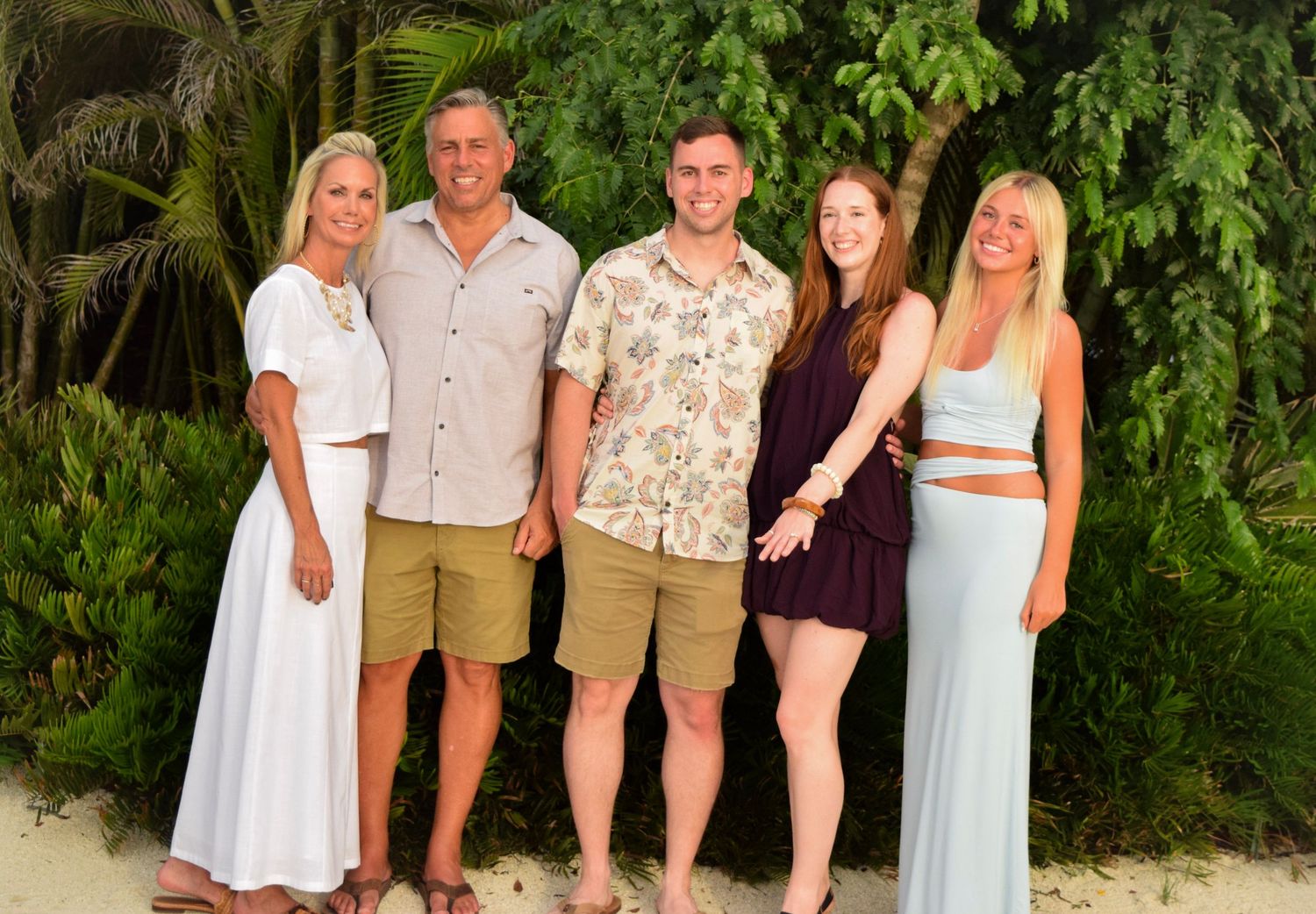 Five people posing together on a tropical beach surrounded by lush green palm trees in a sunny outdoor setting.