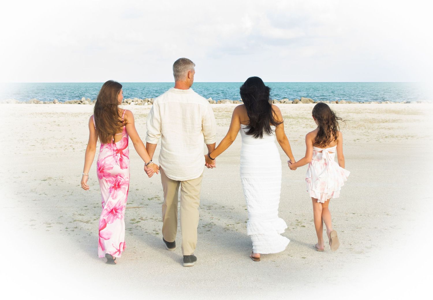Family of four holding hands walking on a sunny beach, dressed in white and pink outfits.