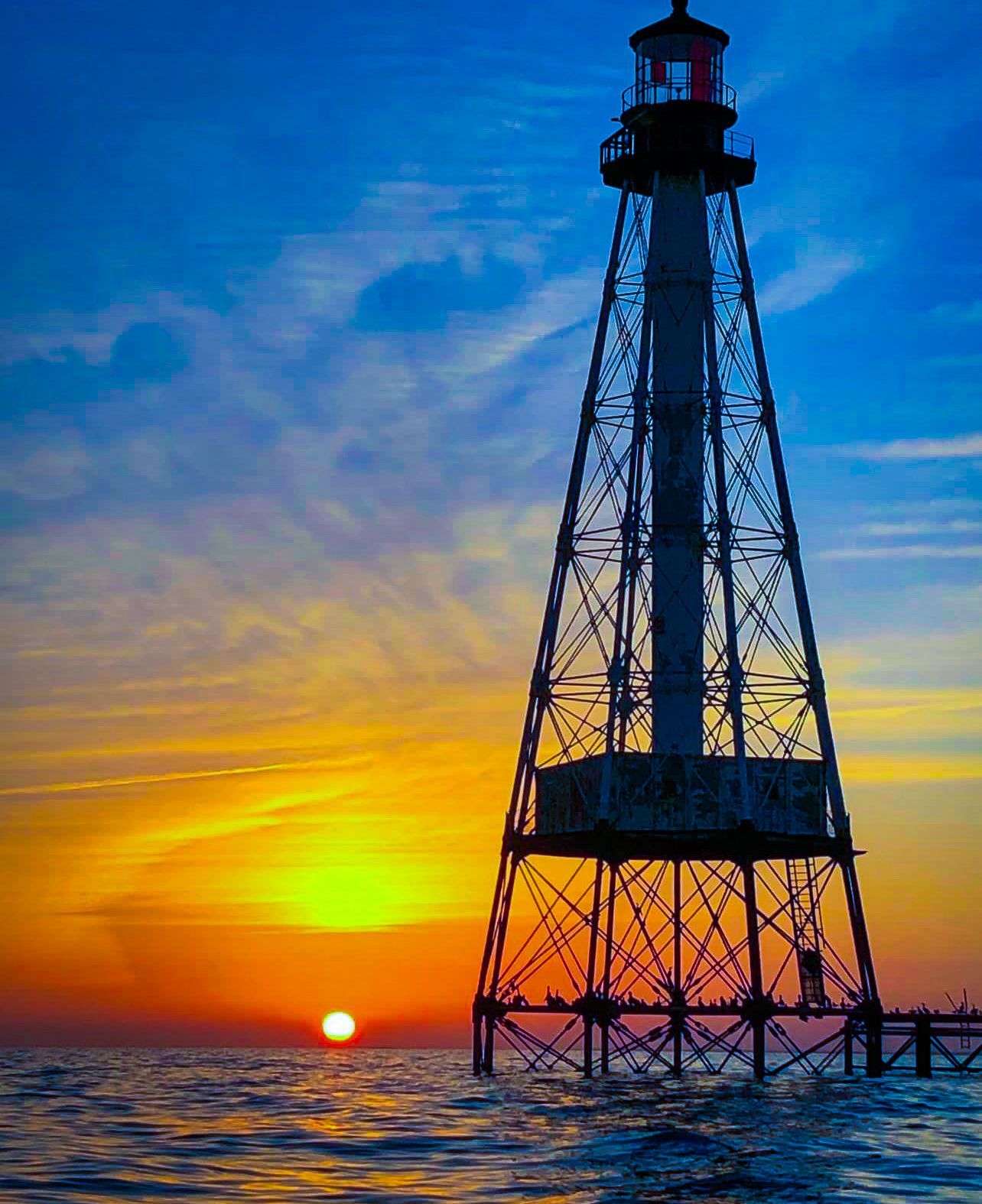A silhouetted offshore lighthouse stands against a vibrant sunset over calm ocean waters with vivid blue and orange skies.
