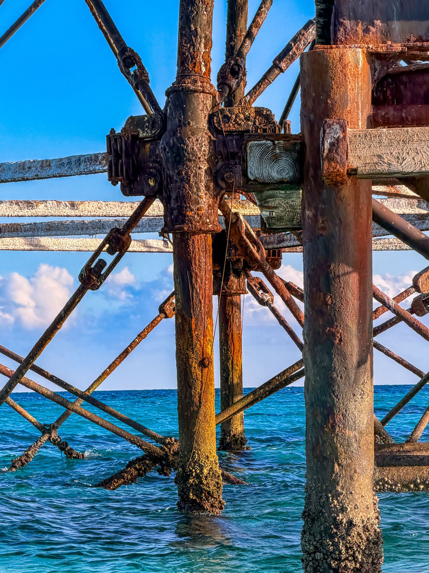 Rusty metal lighthouse structure with chains and corroded poles standing over turquoise ocean water under a blue sky.