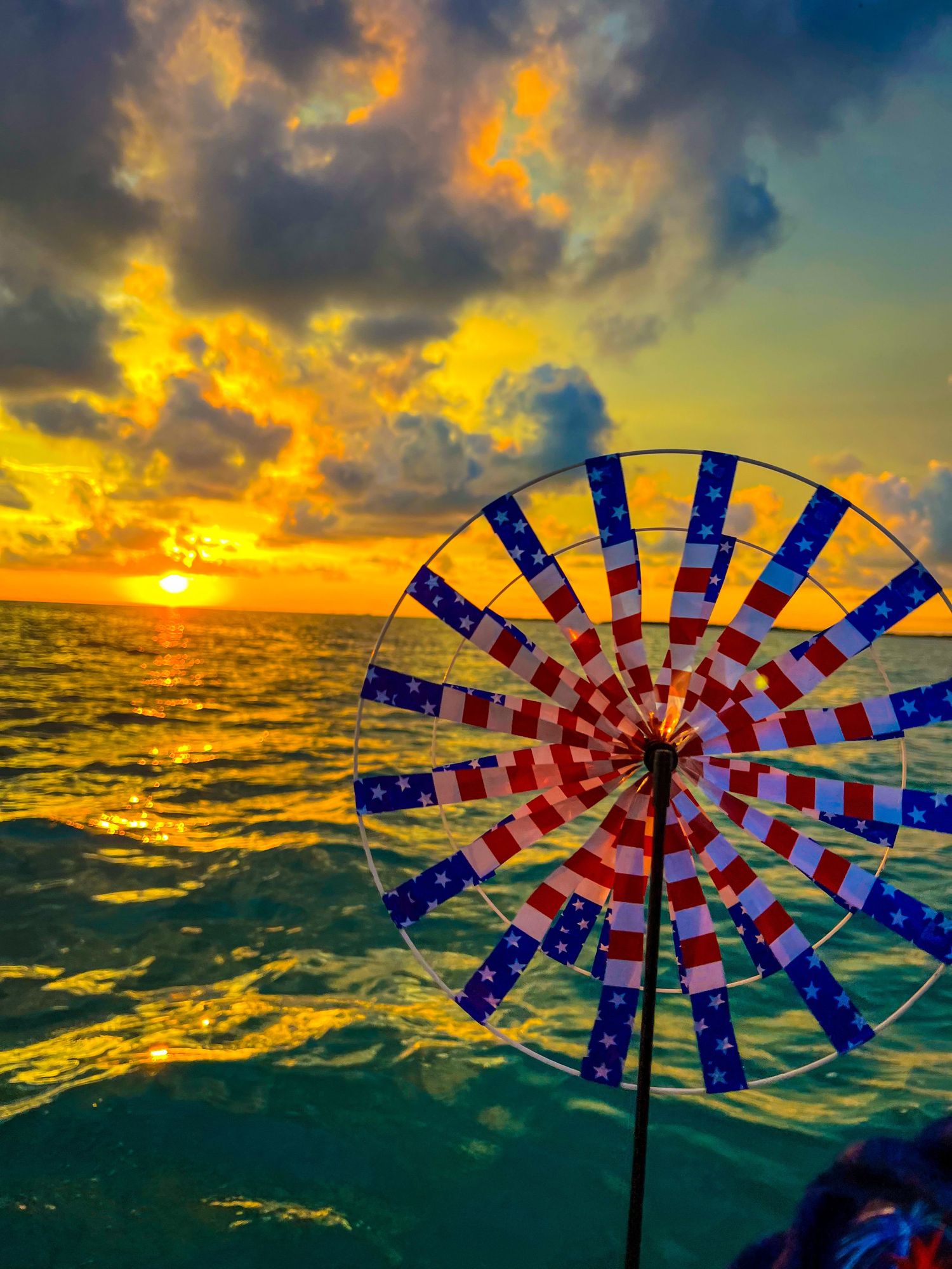 American flag pinwheel spinning against a vibrant ocean sunset with golden clouds and shimmering blue-green waves.