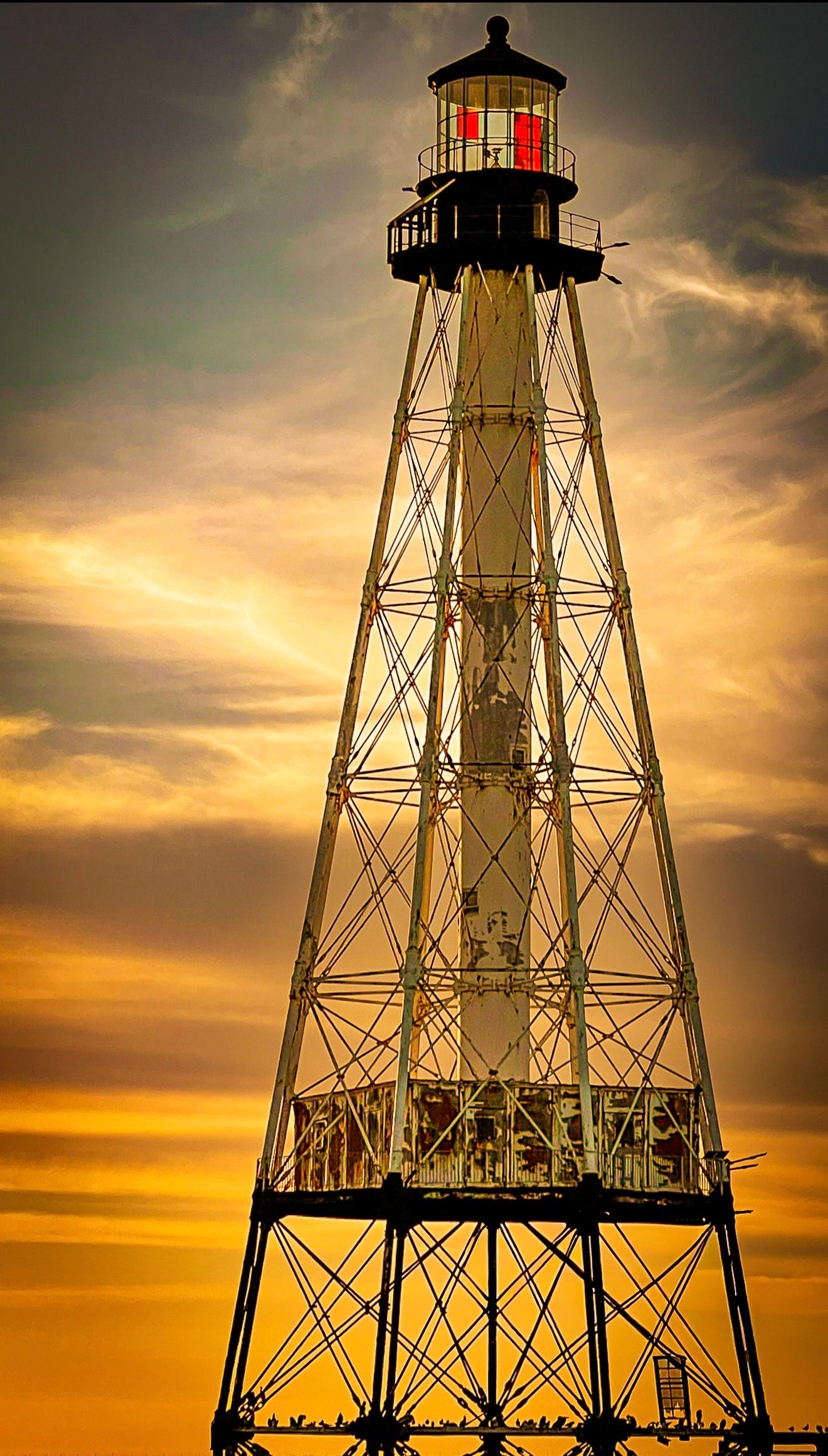 Tall iron lighthouse tower silhouetted against a dramatic golden sunset sky with glowing red light at top.