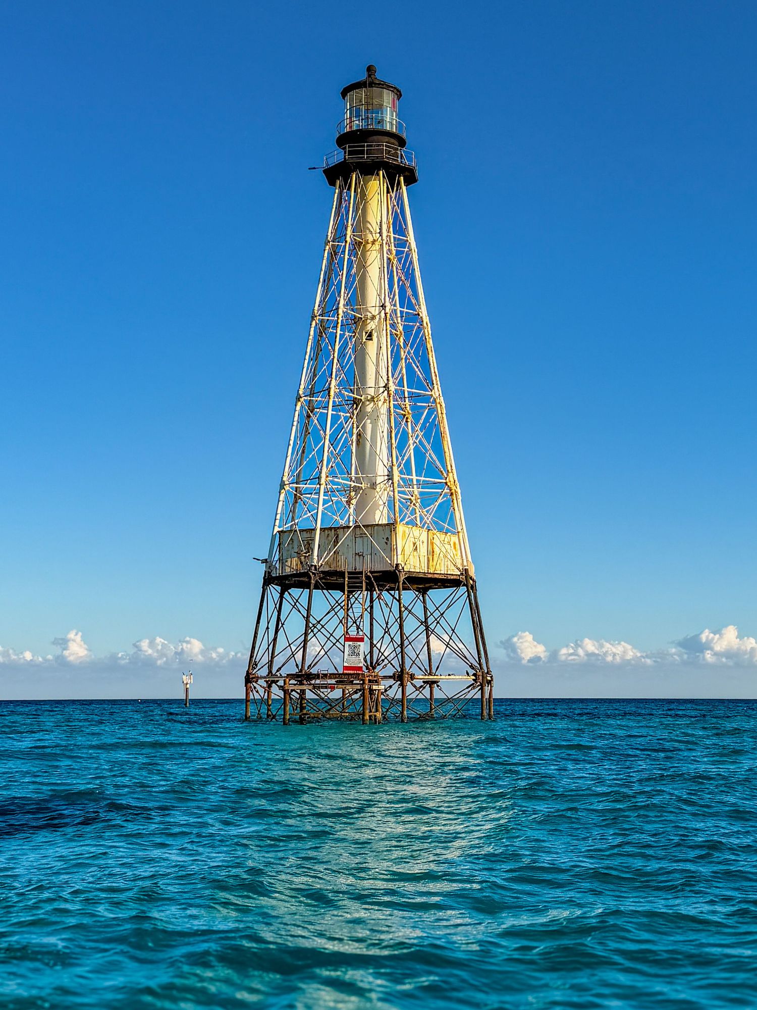 A weathered iron pile lighthouse stands in turquoise ocean waters under a clear blue sky in the Florida Keys.