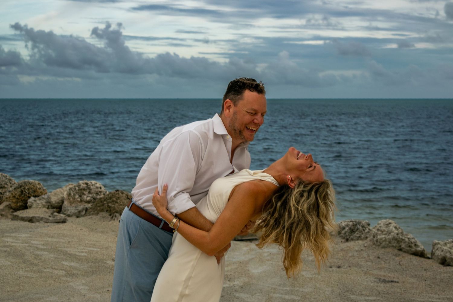A smiling couple in a romantic dip pose on a rocky beach, with the woman leaning back and the ocean and cloudy sky behind them.