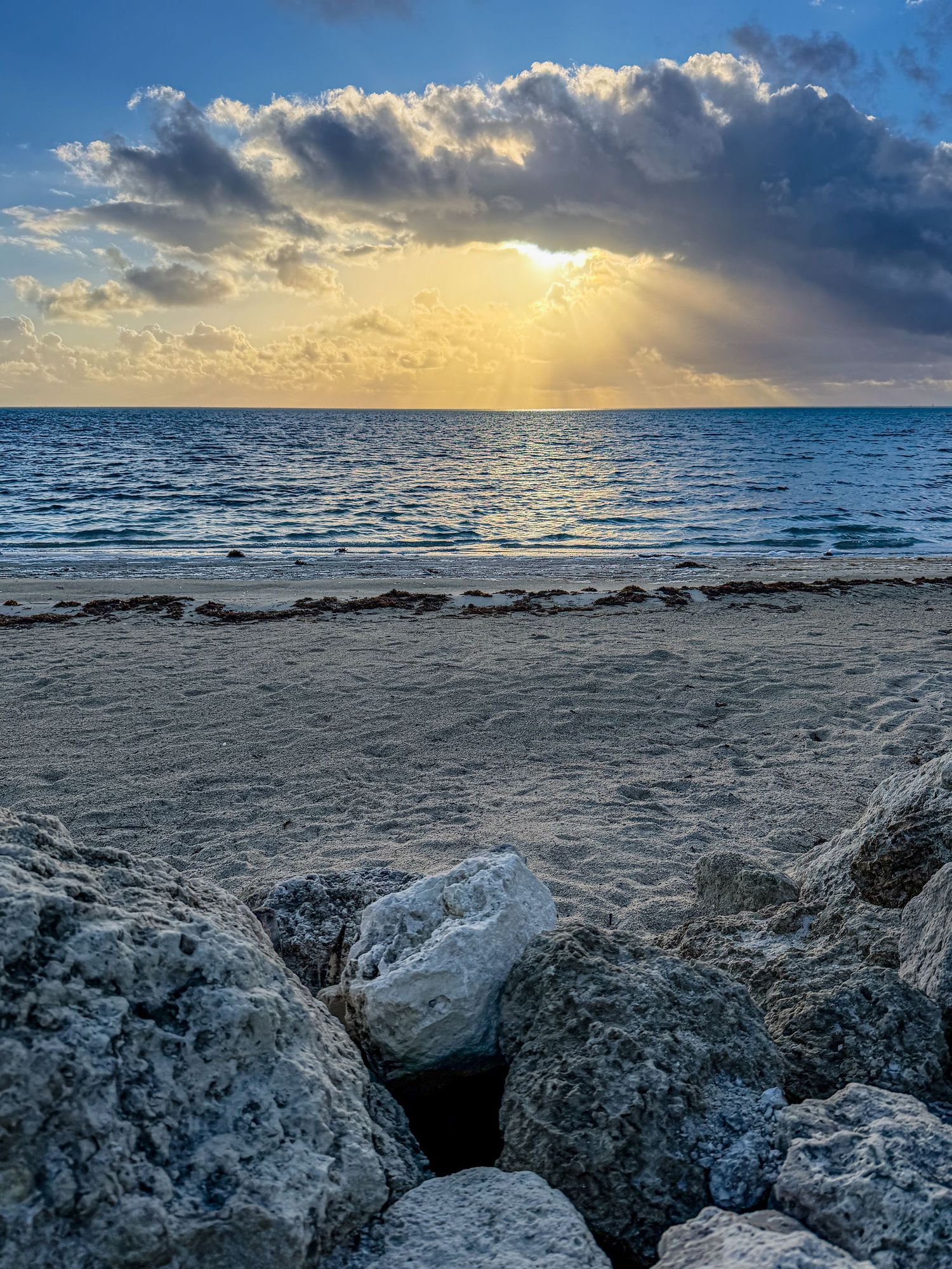 Golden sunset rays breaking through clouds over rocky beach shoreline with calm ocean waves.