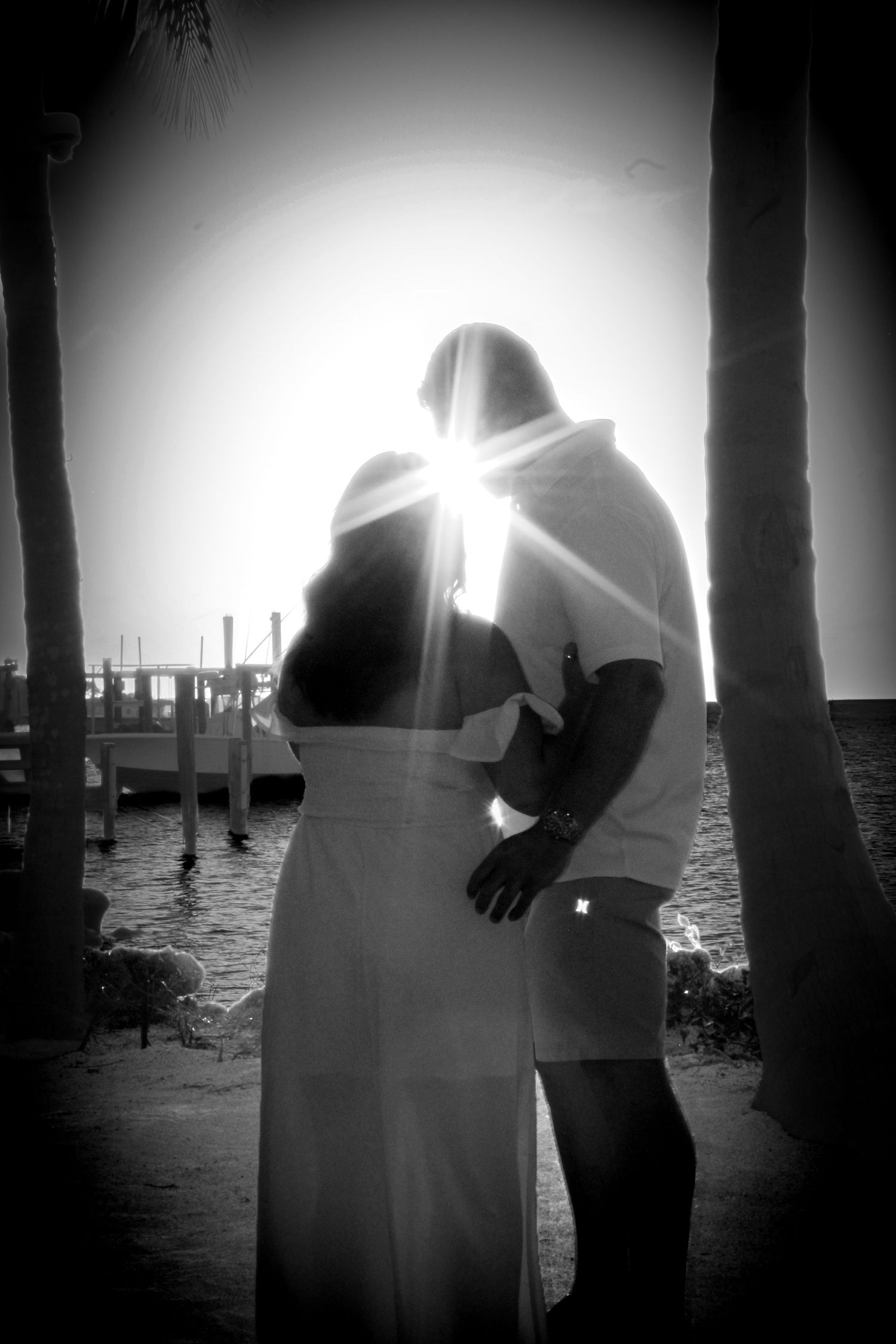 Romantic black and white photo of a couple embracing near palm trees with sunburst effect by a tropical waterfront.