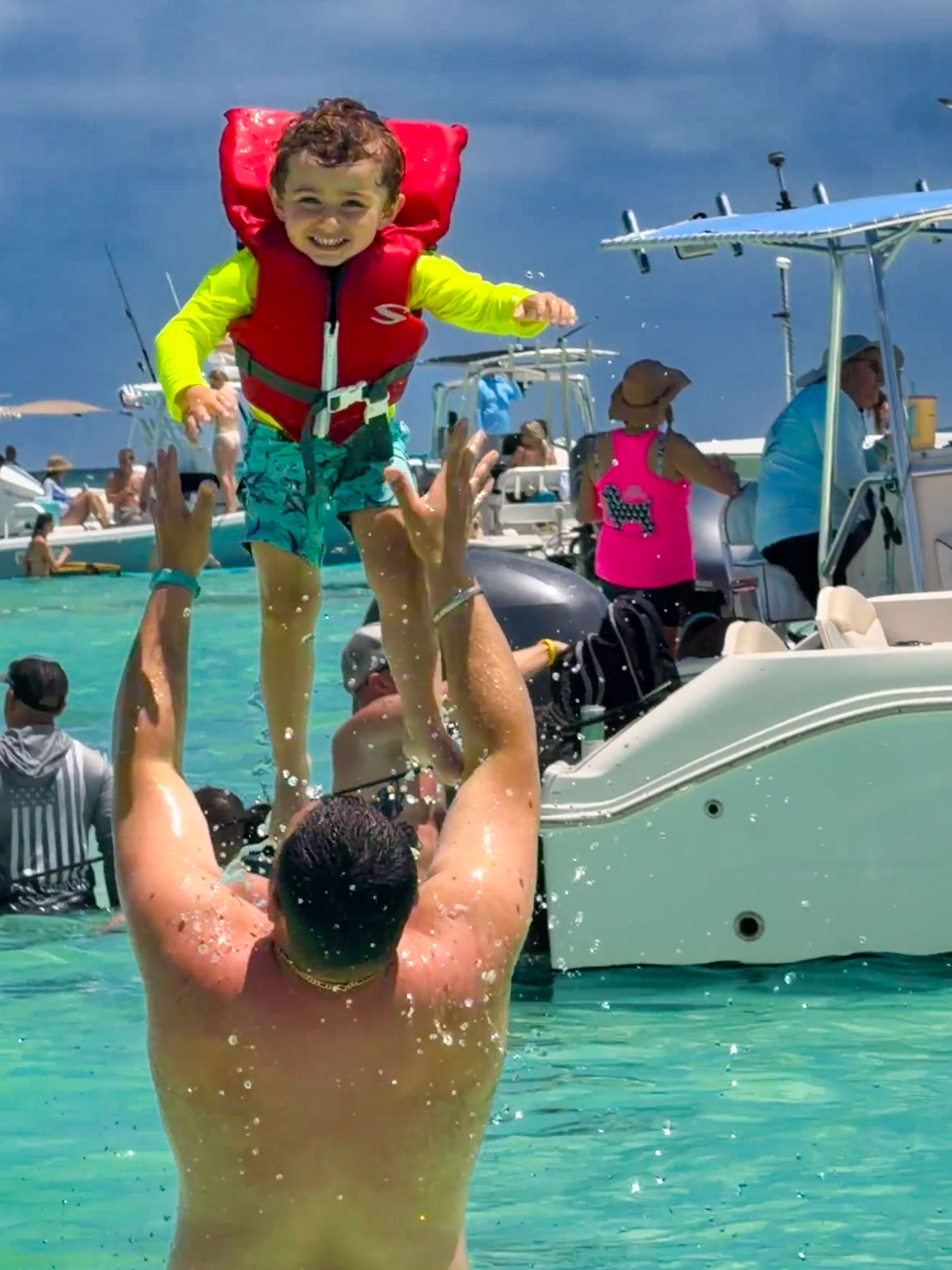 A man tosses a young child wearing a red life jacket into the air in turquoise ocean waters near boats.